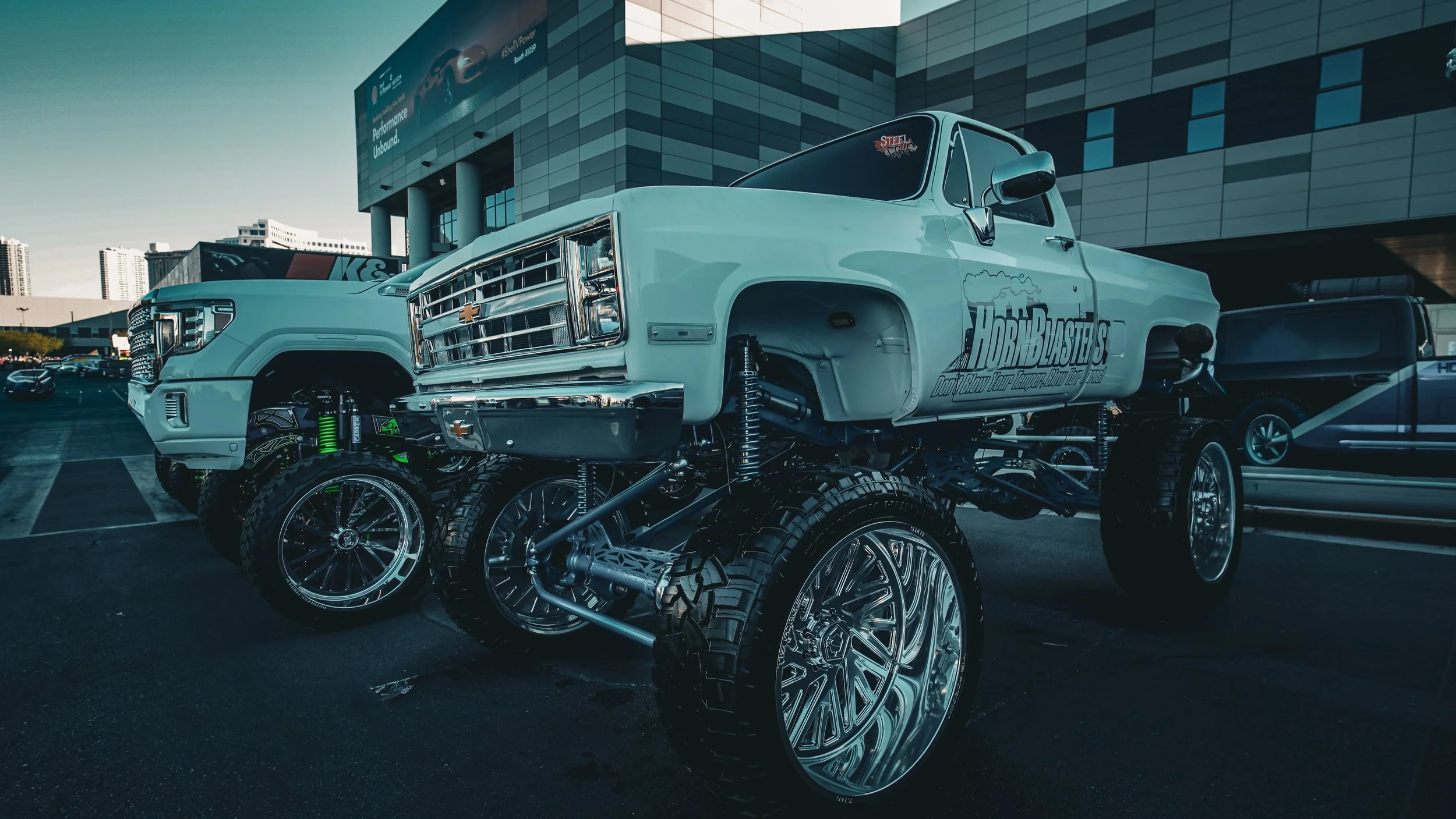A large customized pickup truck with silver body paint and big chrome wheels, parked in a lot near a modern building.
