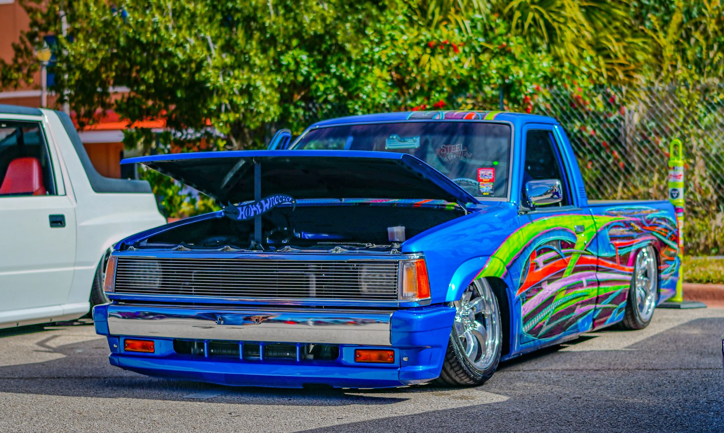 A customized blue pickup truck with a vibrant, colorful flame design on the side, lowered stance, chrome wheels, and an open hood, parked on the street during daytime.