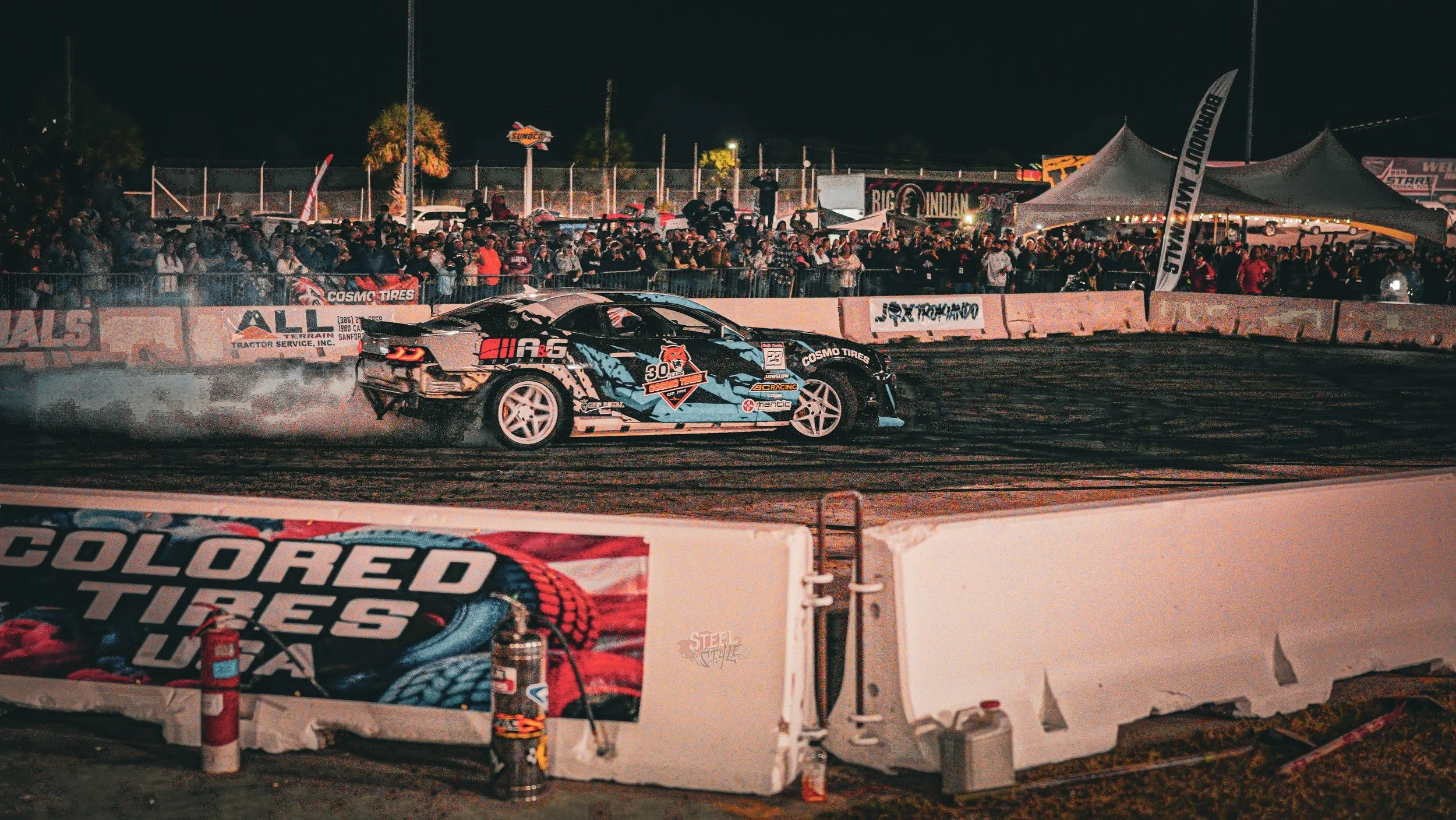 A racing car performs a drift on a dirt track at night during a motorsport event, with a crowd of spectators in the background and banners displaying tire brands and event logos.