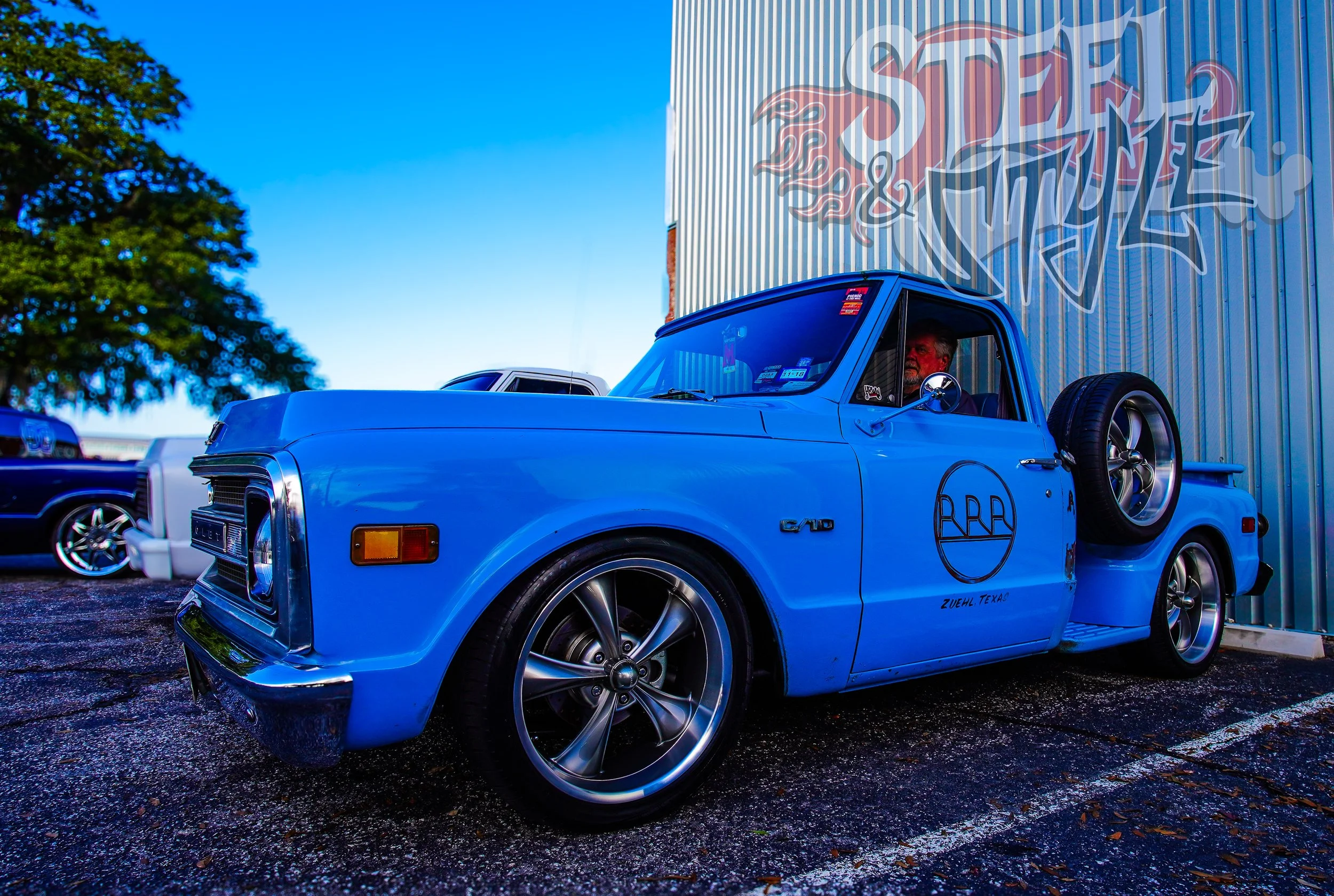 Blue vintage pickup truck parked outside a building with graffiti art. The truck has custom wheels, a spare tire mounted on the side, and a logo on the door reading 'Zuehl, Texas.' There are other vehicles and a large tree in the background under a c
