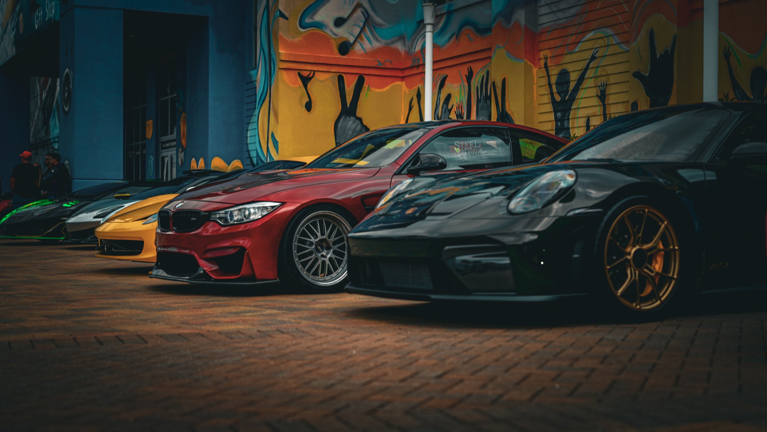 Multiple sports cars parked in front of a colorful graffiti wall, including a black car with gold wheels, a red BMW, a yellow car, and a black car with green accents.