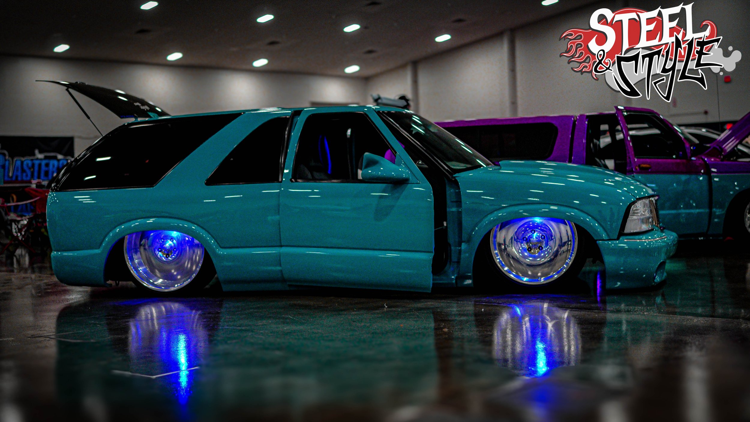 A customized, teal-colored, lowrider car with illuminated blue wheels on display at an indoor car show, with the 'Steel & Style' logo in the top right corner.
