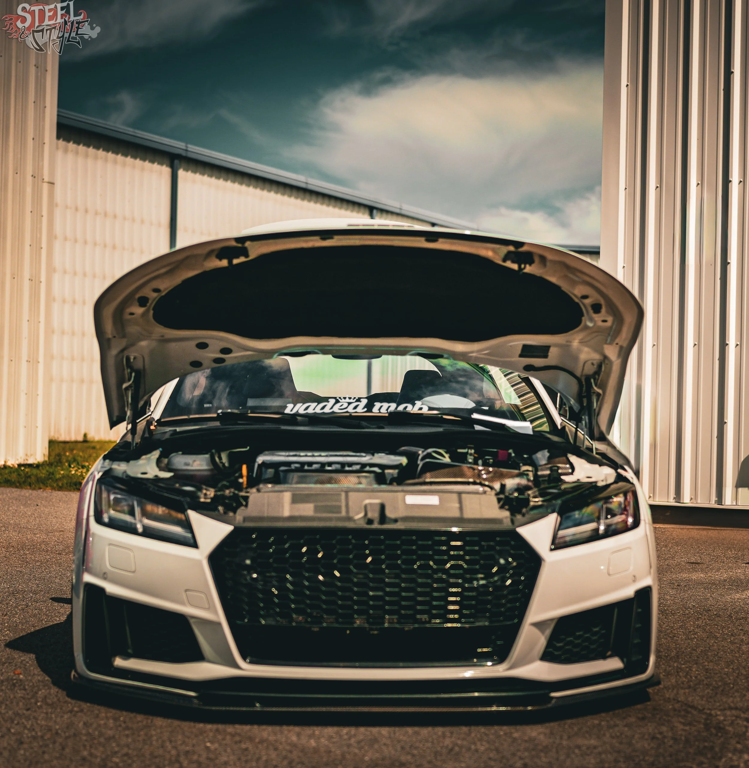 Front view of a white car with its hood open, parked outside near corrugated metal walls, under a cloudy sky.