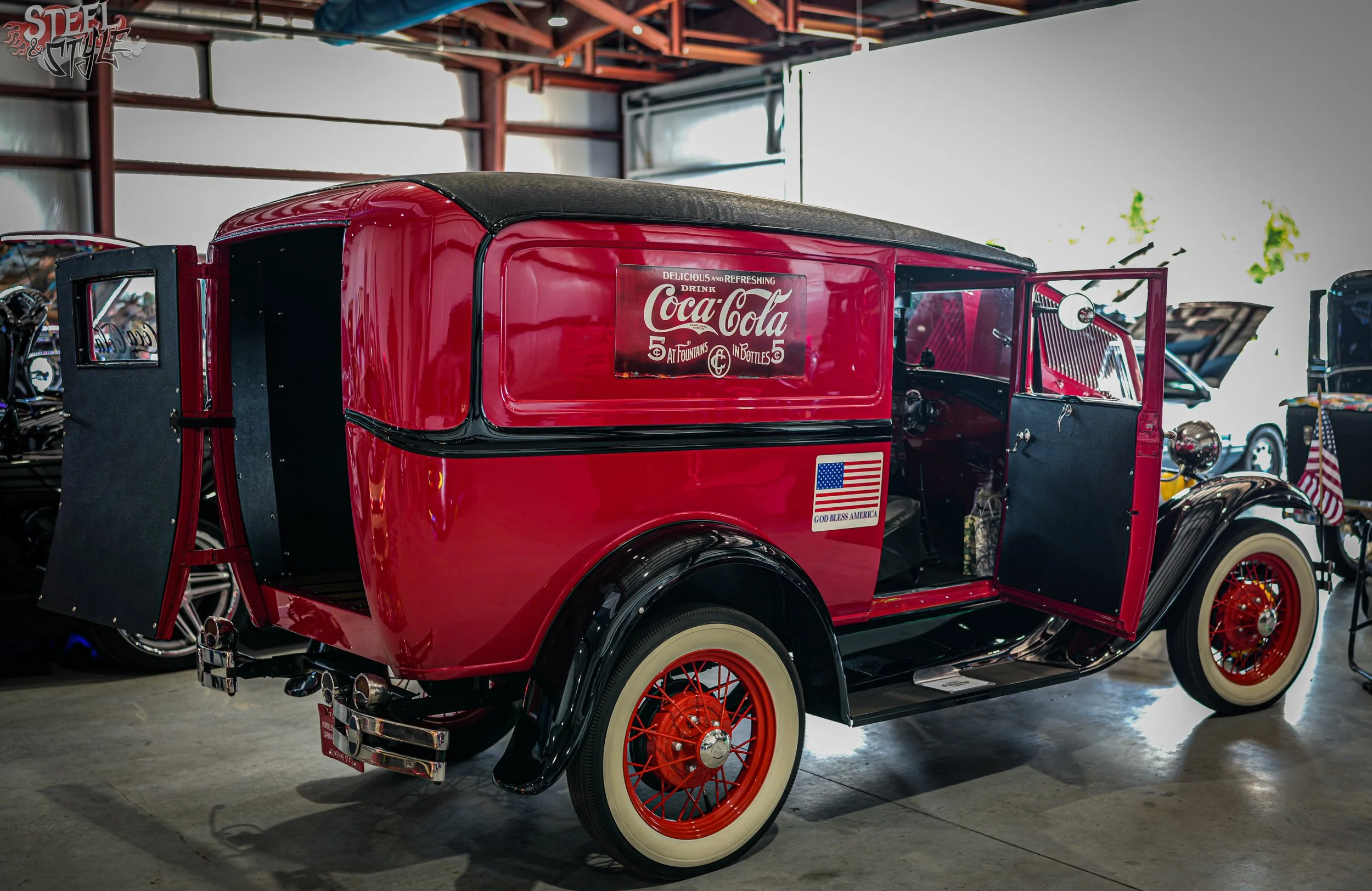 A vintage red Coca-Cola delivery truck with white wheels and whitewall tires, displayed indoors with a wooden ceiling and other vintage vehicles visible in the background.