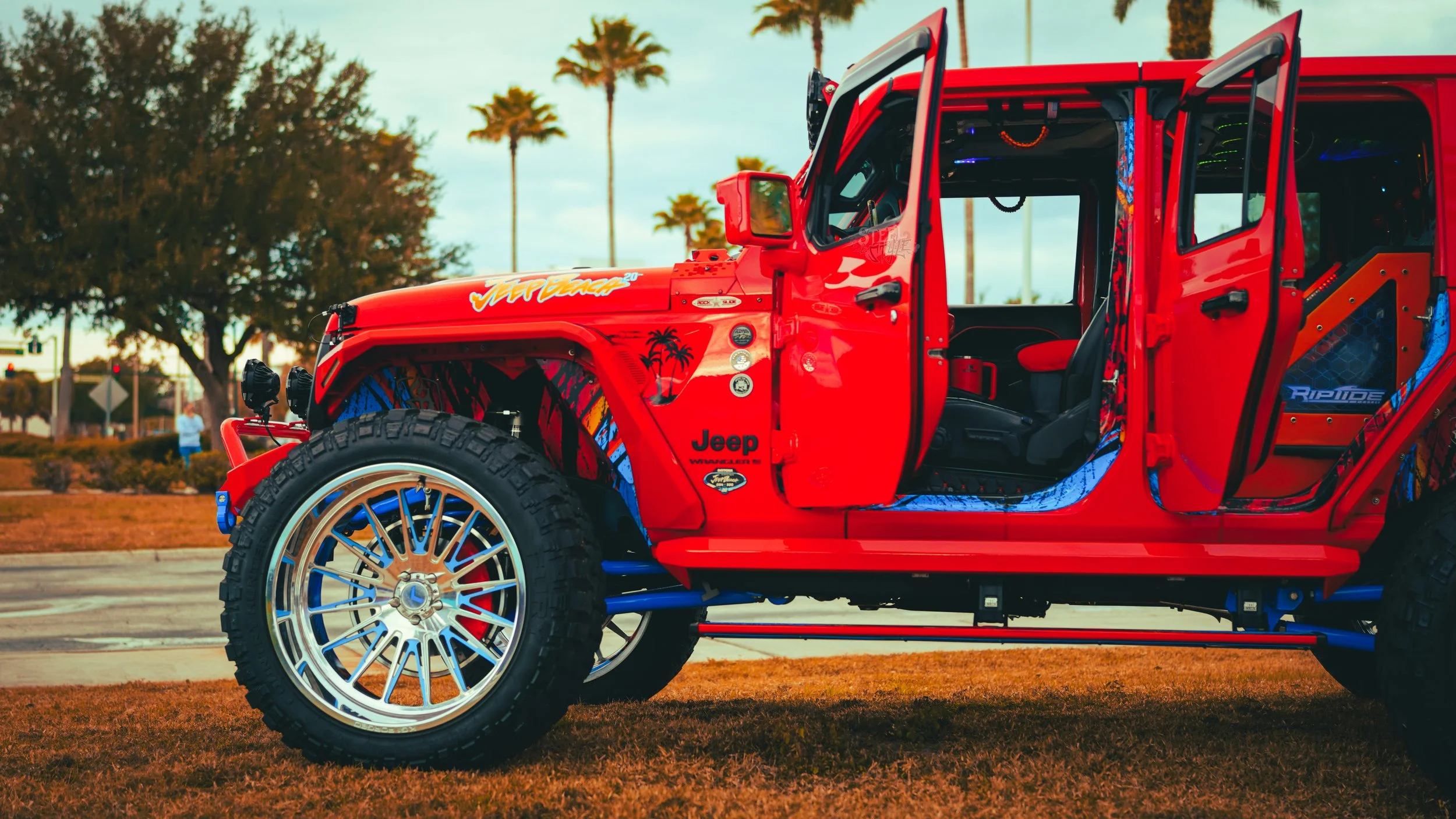 A red Jeep Wrangler with open doors parked on grass, featuring custom wheels, decals, and a modified interior, with trees and a street in the background.