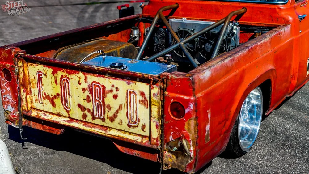 Vintage red Ford pickup truck with rust and scratches, showing the rear with a rusty, weathered Ford sign, custom wheels, and an engine exposed in the bed.