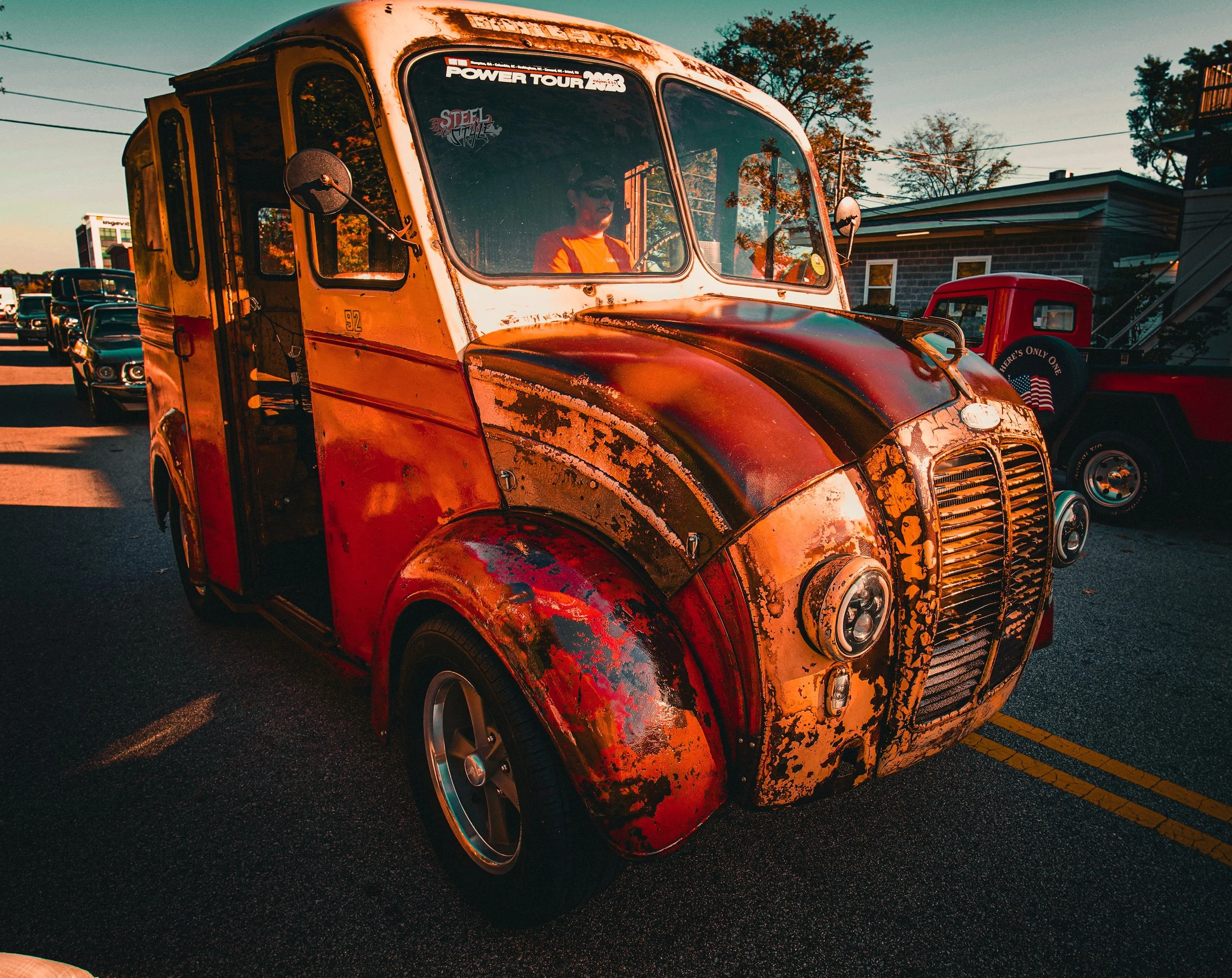 A vintage, rusty orange and black delivery vehicle with a weathered appearance, parked on a street during sunset, with other classic cars and vintage trucks visible in the background.