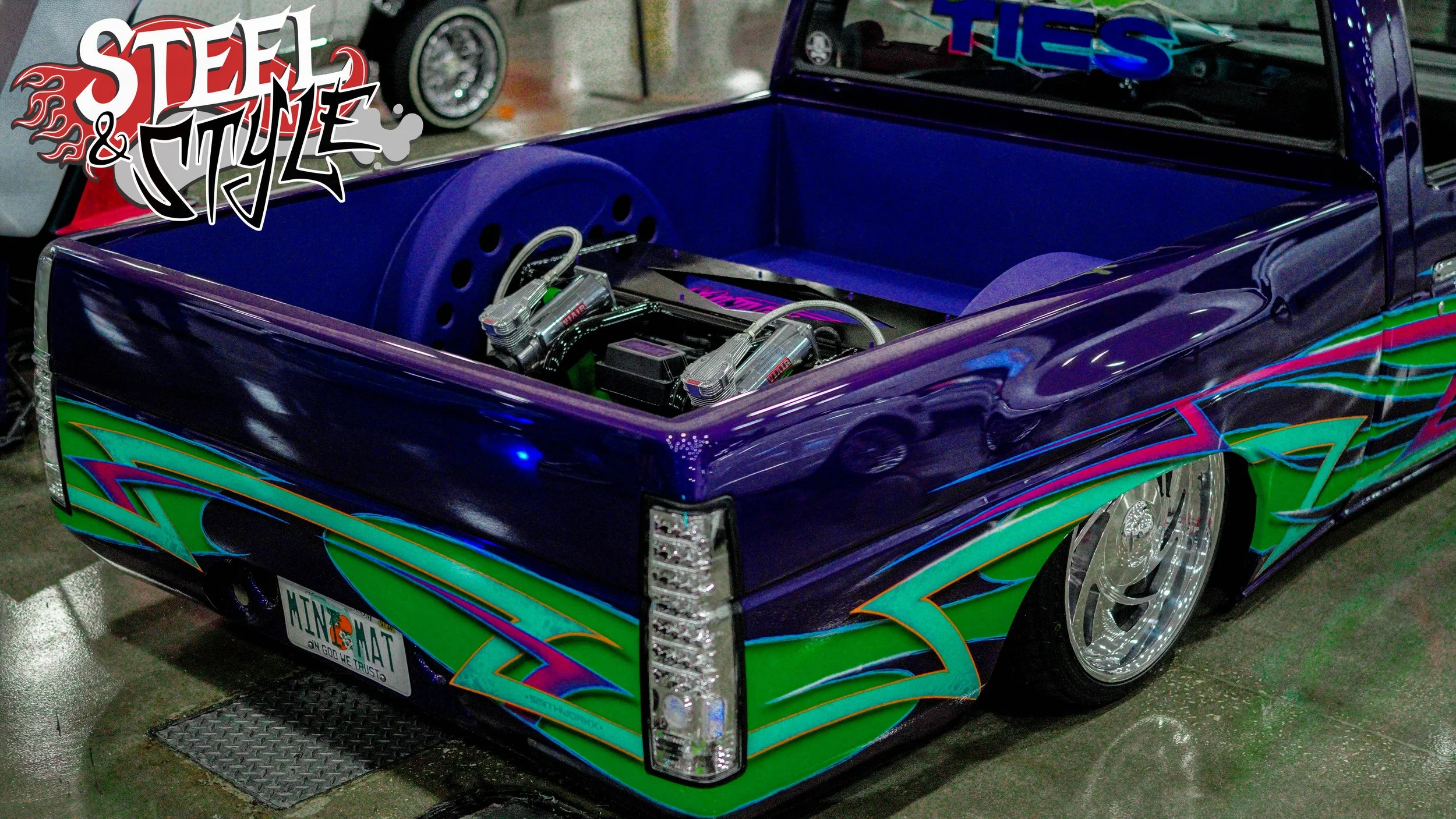 Custom lowrider pickup truck with a vibrant, colorful graphic paint job in green, purple, and blue, featuring a stripped-down bed with visible engine components, chrome wheels, and a license plate that reads 'HIN MAT'.