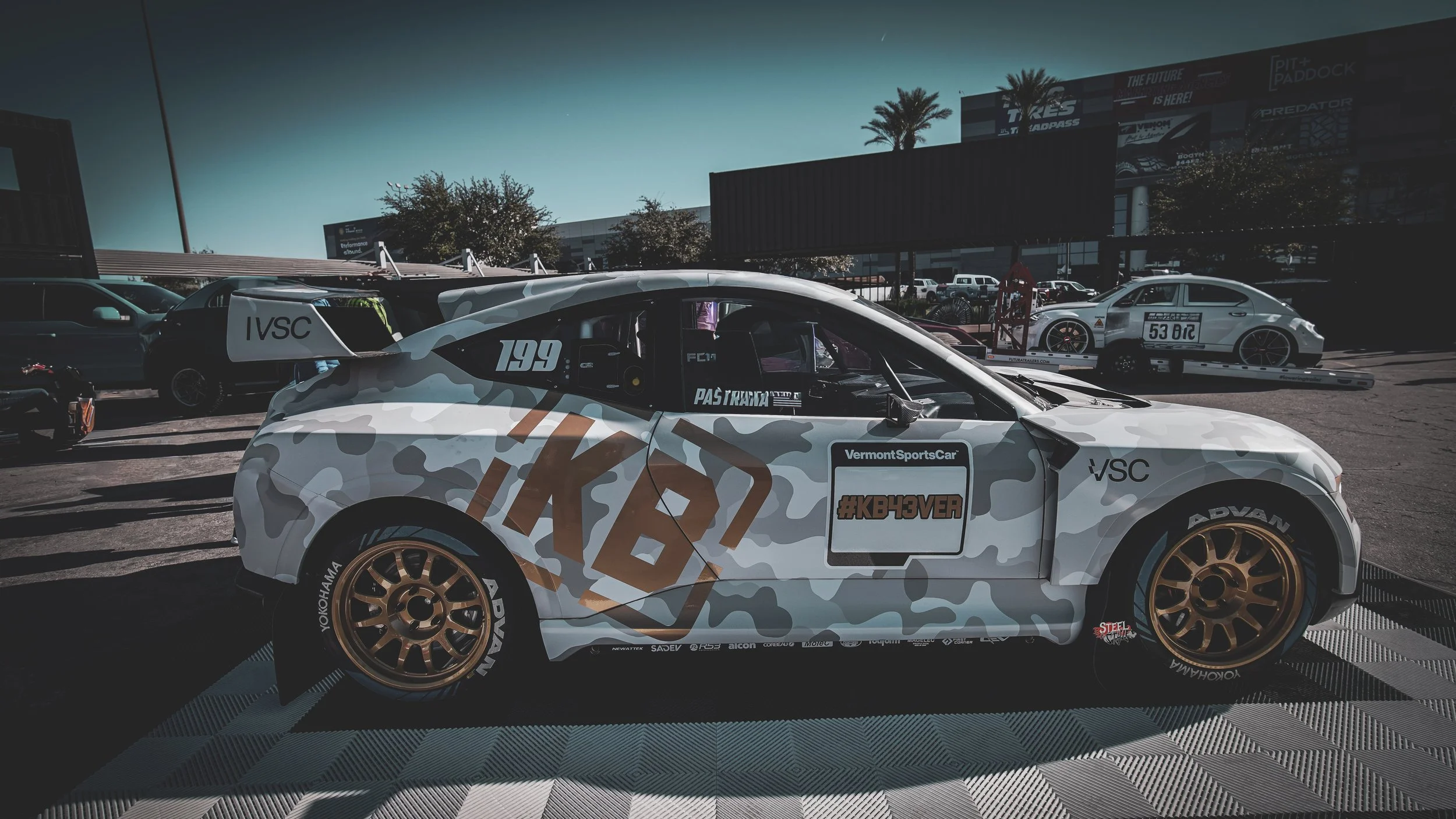 A race car with a camouflage pattern and gold wheels, parked outdoors in a paddock area with other racing vehicles and trailers visible in the background.