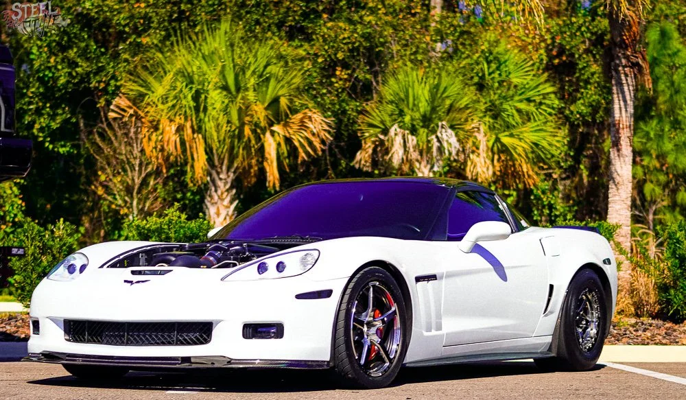 White sports car parked outdoors with a tropical background of palm trees and lush greenery.