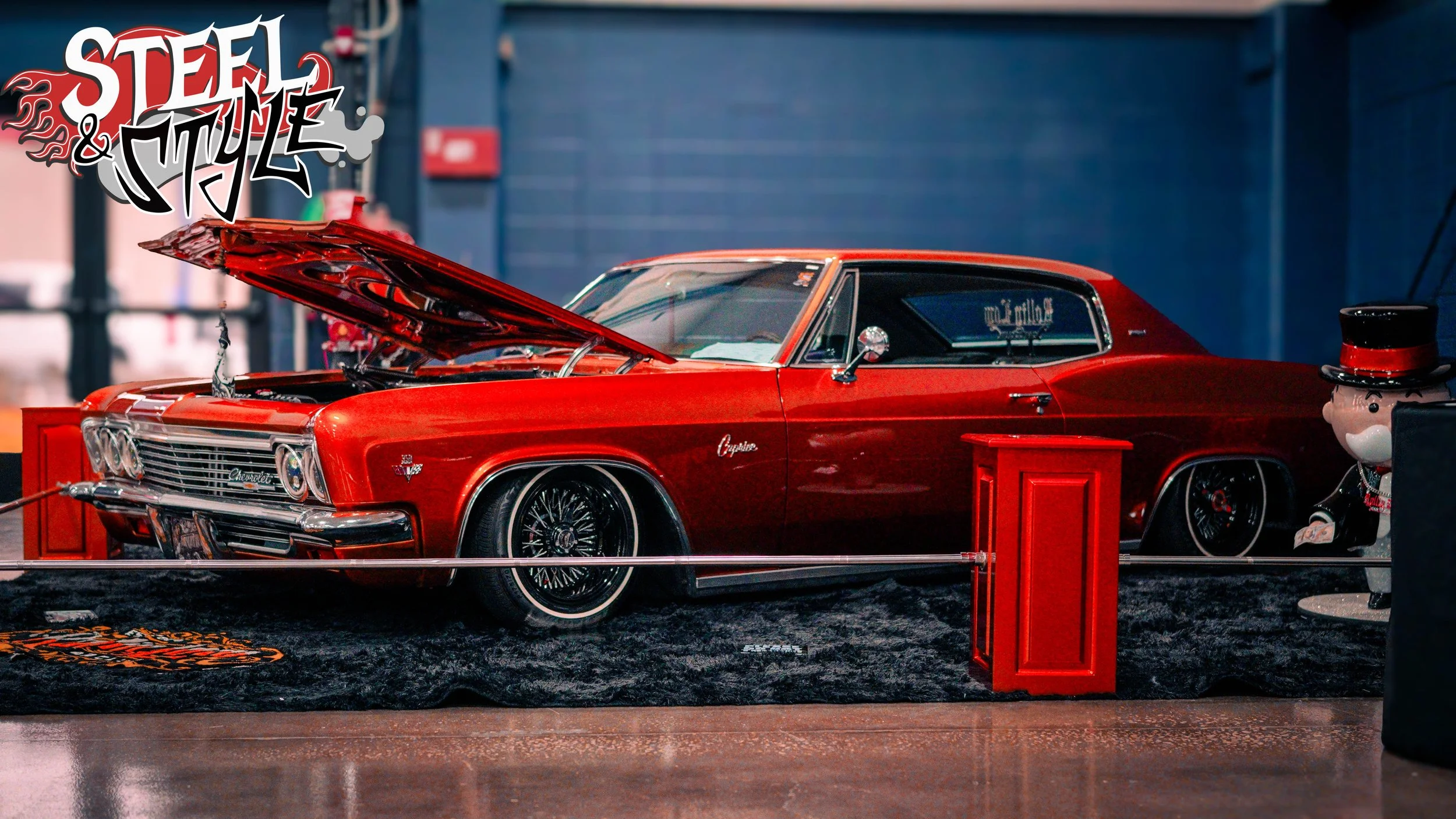 A red vintage Chevrolet Impala with its hood open displayed at an indoor car show, decorated with a 'Steel & Style' logo in the top left corner, and a small snowman figure with a top hat on the right side.