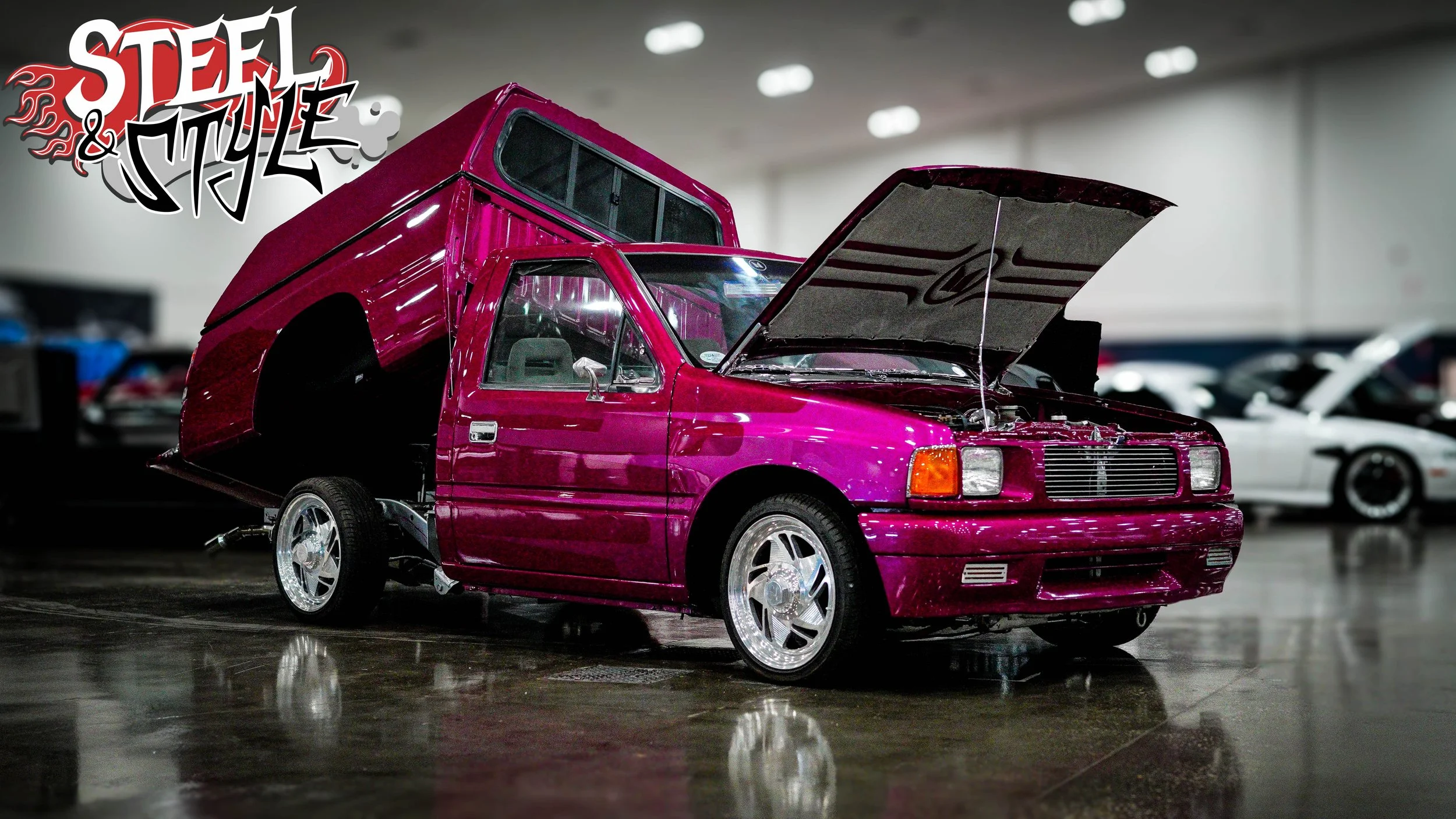 Modified pink pickup truck with the hood open and a raised bed in an indoor showroom.
