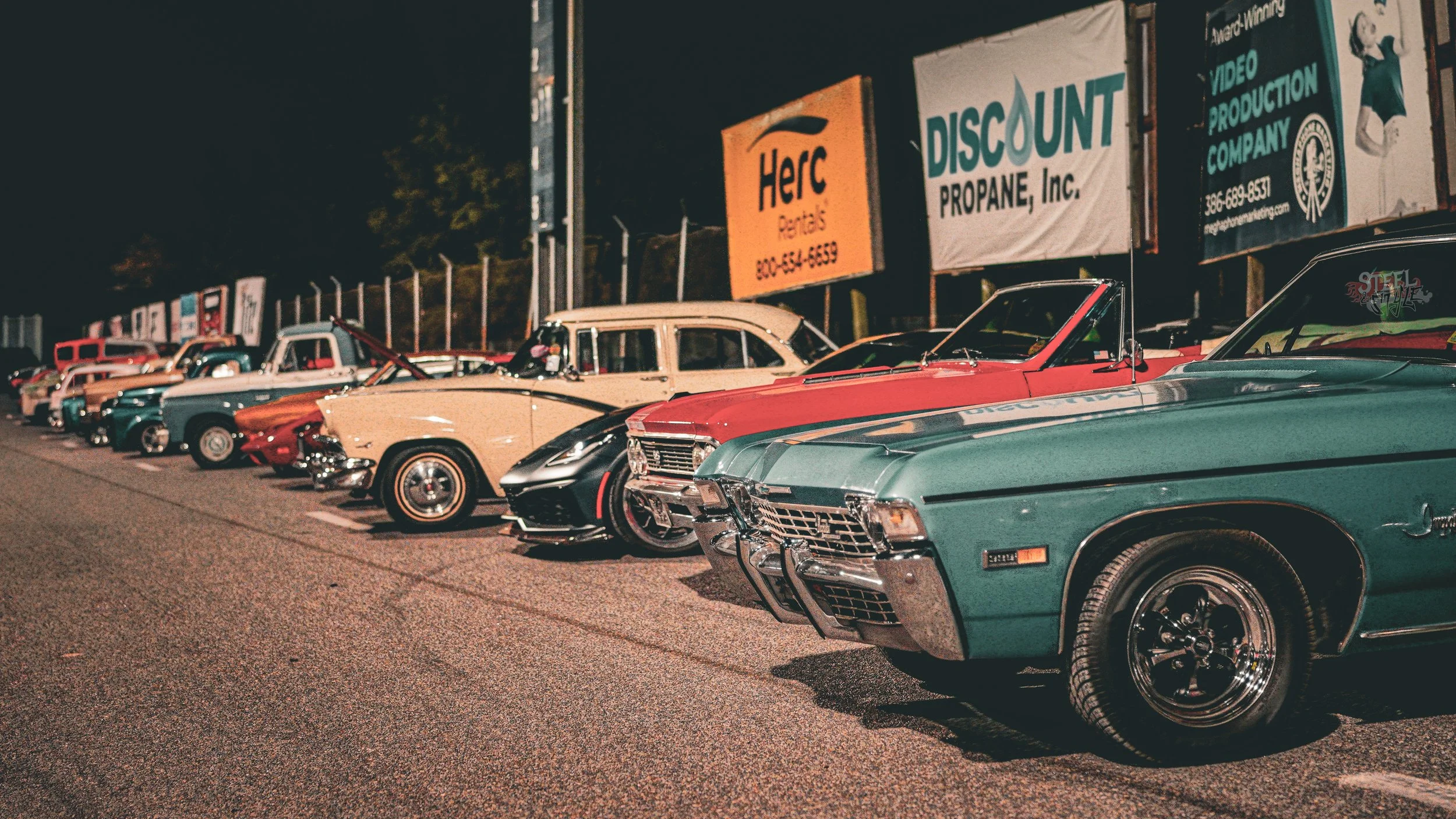 A lineup of vintage and classic cars parked side by side at night, with advertising signs in the background.