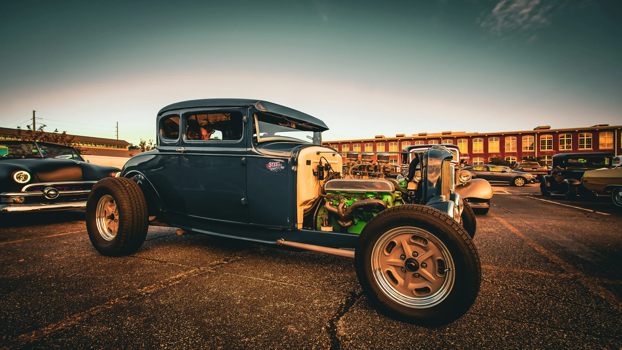Vintage car on display in a parking lot during the evening, with other classic cars and a building in the background.