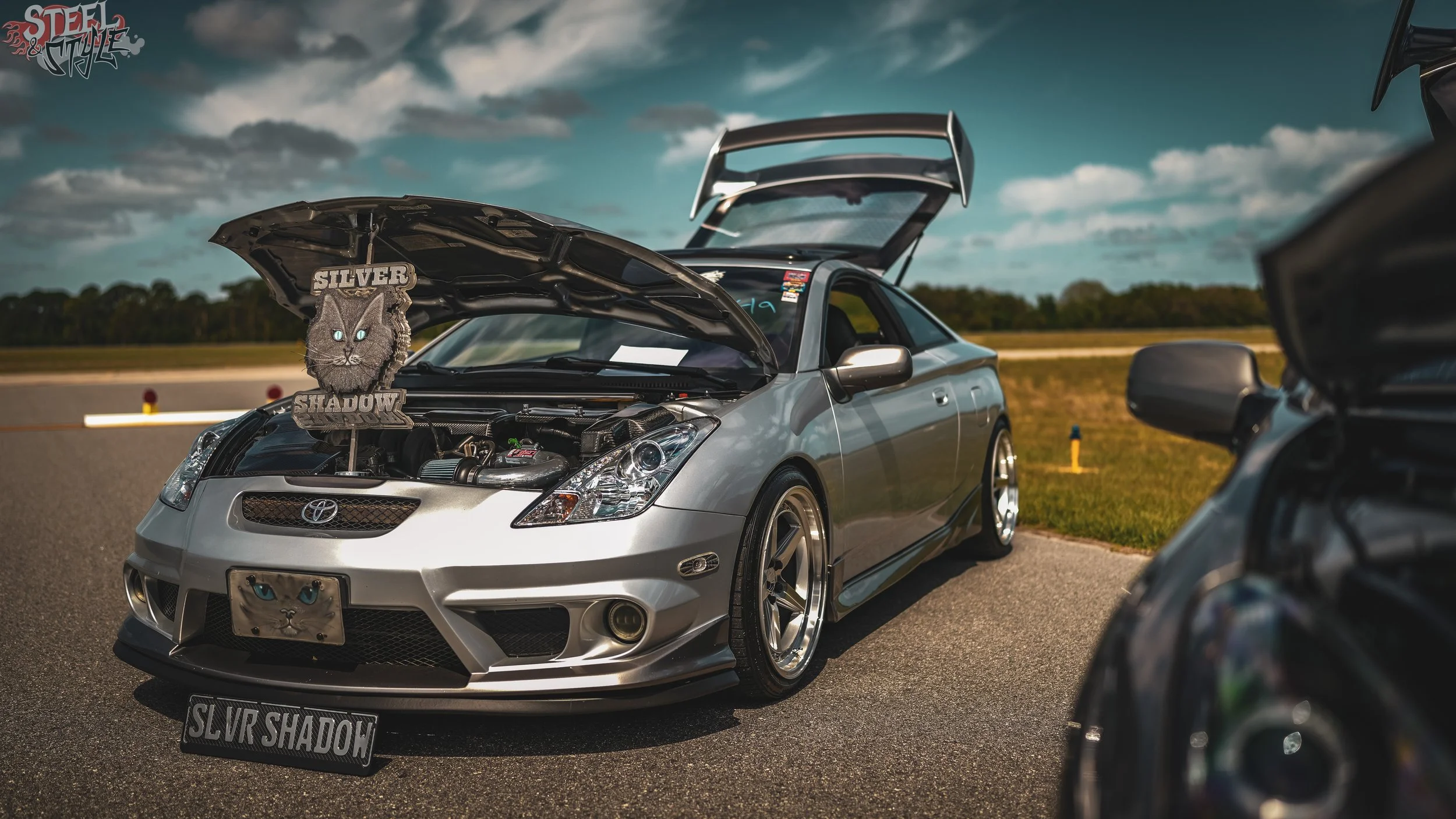 A silver sports car with its hood and trunk open parked on a race track, featuring a custom badge with a black cat face and the words 'Silver Shadow' on it. There is a blue sky with clouds and trees in the background.