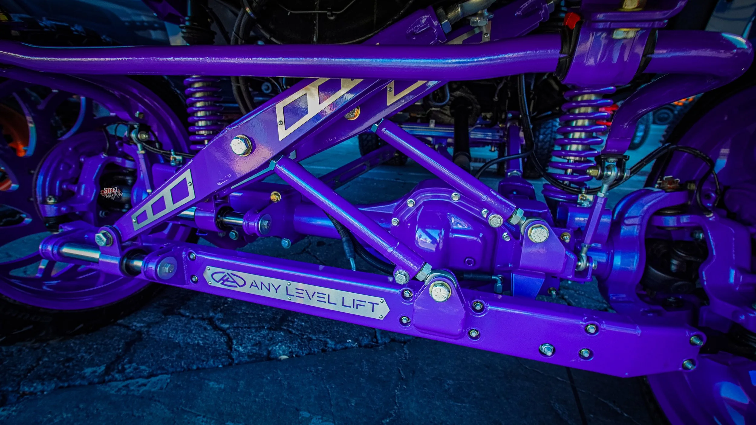 Close-up of a purple-painted vehicle suspension with visible mechanical parts, hydraulic components, and labeled 'ANY LEVEL LIFT'