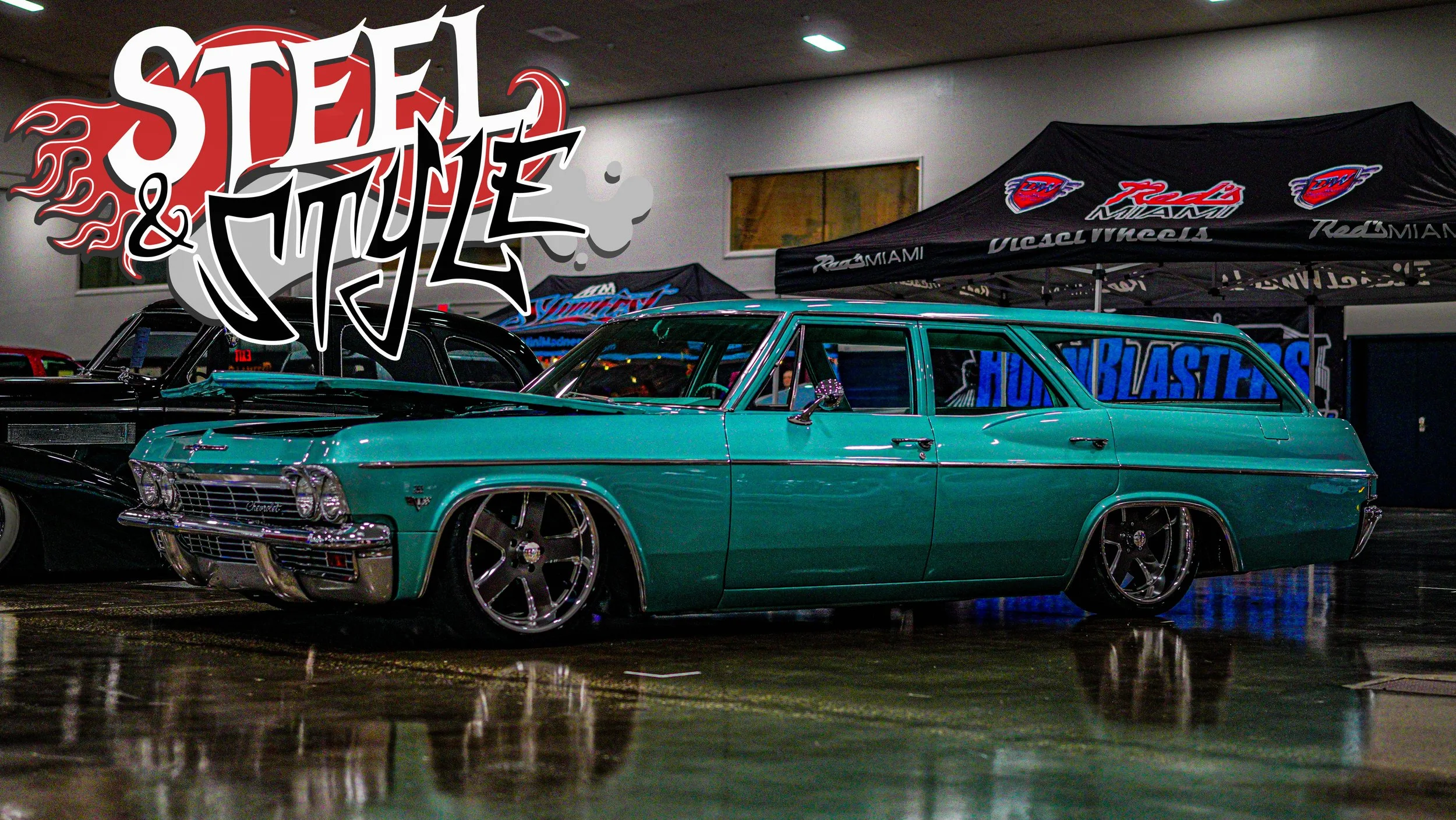 A vintage teal station wagon displayed indoors at a car show, with custom wheels and chrome details, behind a banner that reads 'HUMBLASTERS' and a large graphic text overlayed that says 'Stang & Style' with flames.