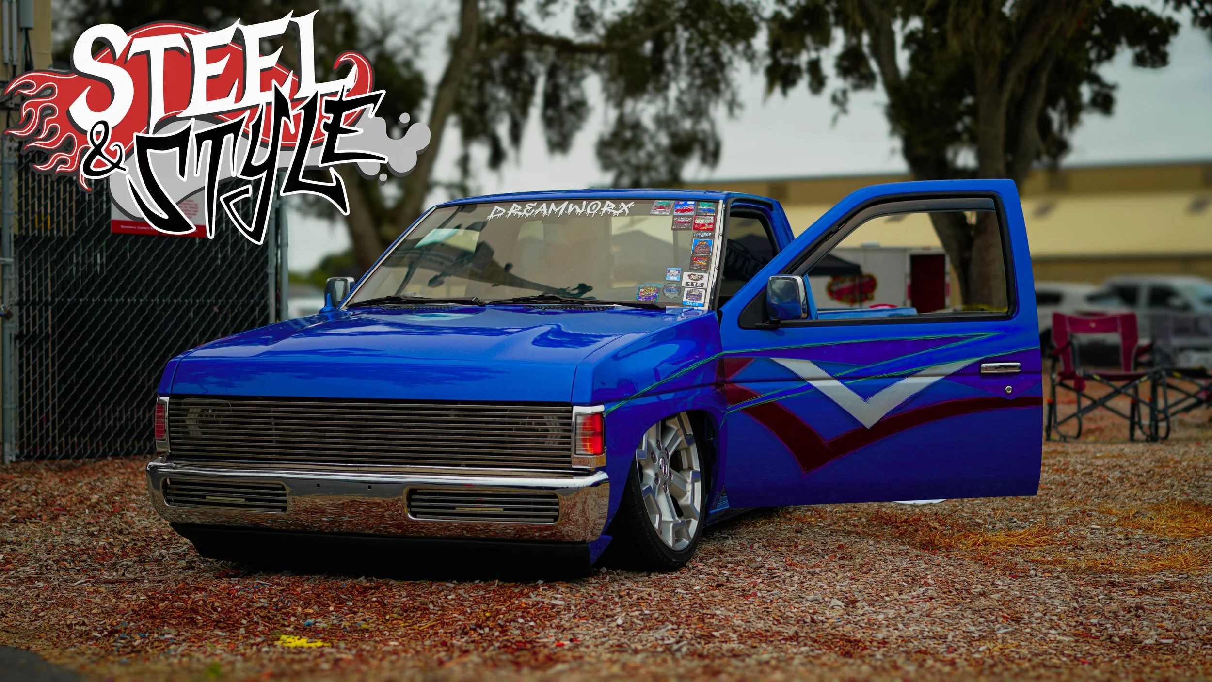 Custom painted blue pickup truck with graphics on side, parked outdoors with open door, at car event or show, with folding chairs and trees in background.