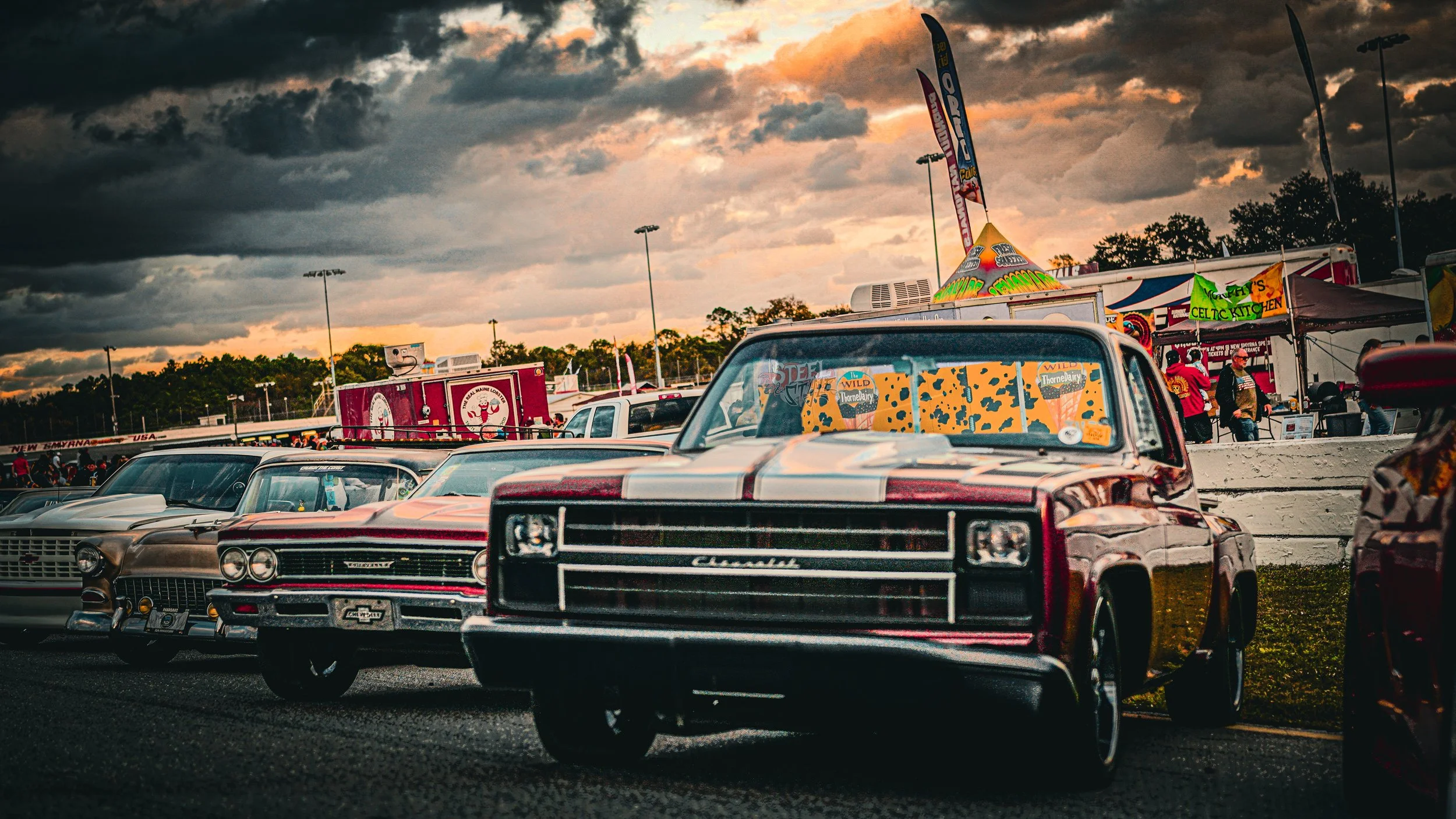 Row of vintage cars parked at an outdoor event during cloudy weather at sunset, with food stalls and tents in the background.