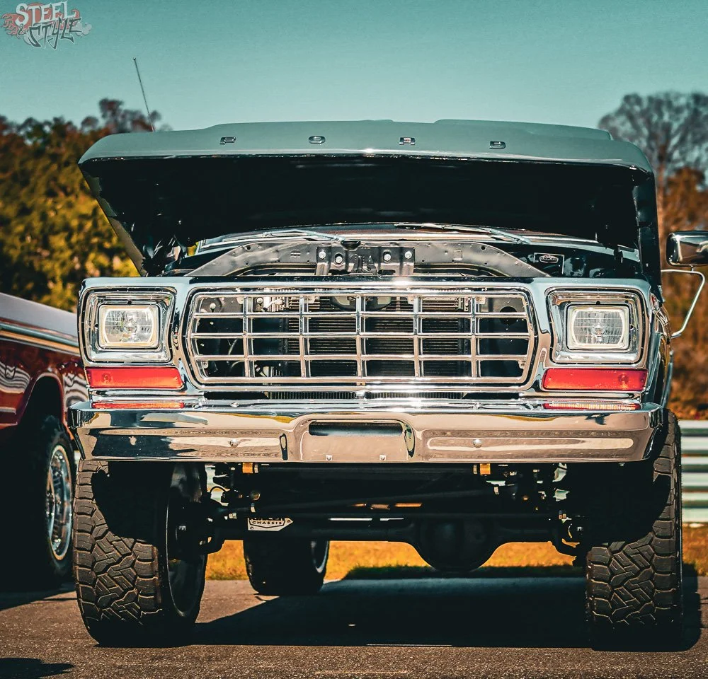Front view of a lifted Ford truck with open hood, chrome grille, rectangular headlights, large off-road tires, parked outdoors during daytime.