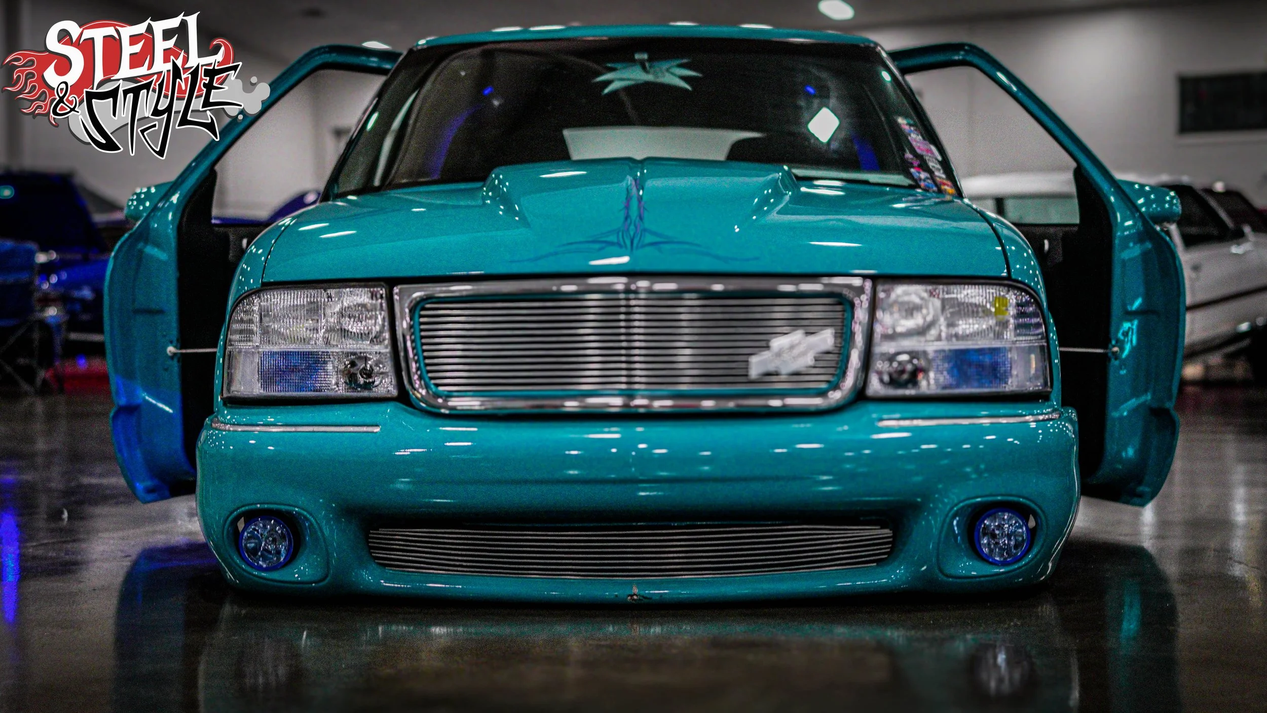 A turquoise car with its front and side doors open, parked indoors with reflections on the polished floor. The car has a custom chrome grille and headlights, with a logo on the front. The background shows other vehicles and a logo that says "Steel & 
