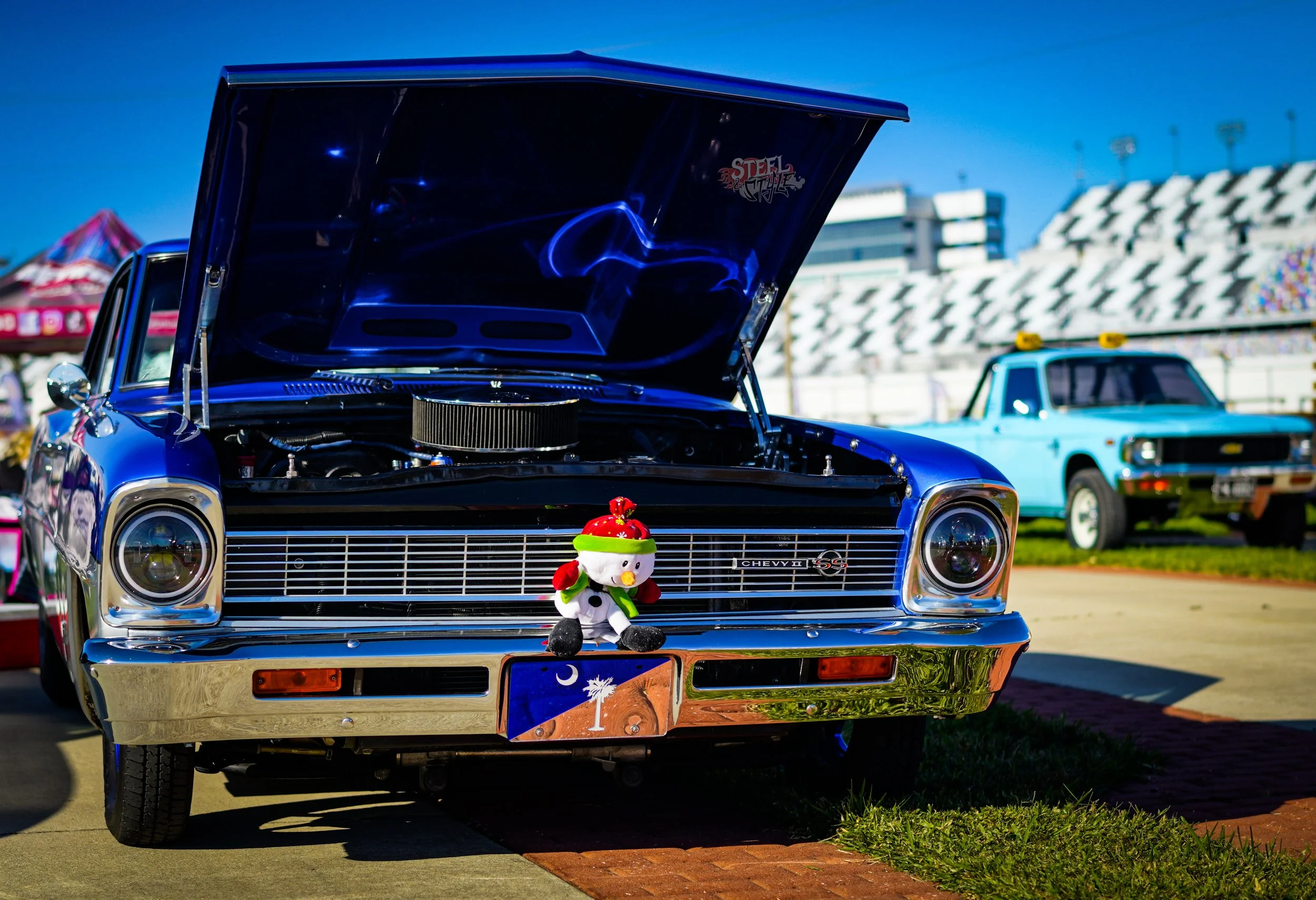 A classic blue Chevrolet car with its hood open, at a car show, with a snowman doll dressed in Christmas attire on the front bumper, and an orange, blue, and white themed license plate. In the background, there is a blue pickup truck and a racetrack 