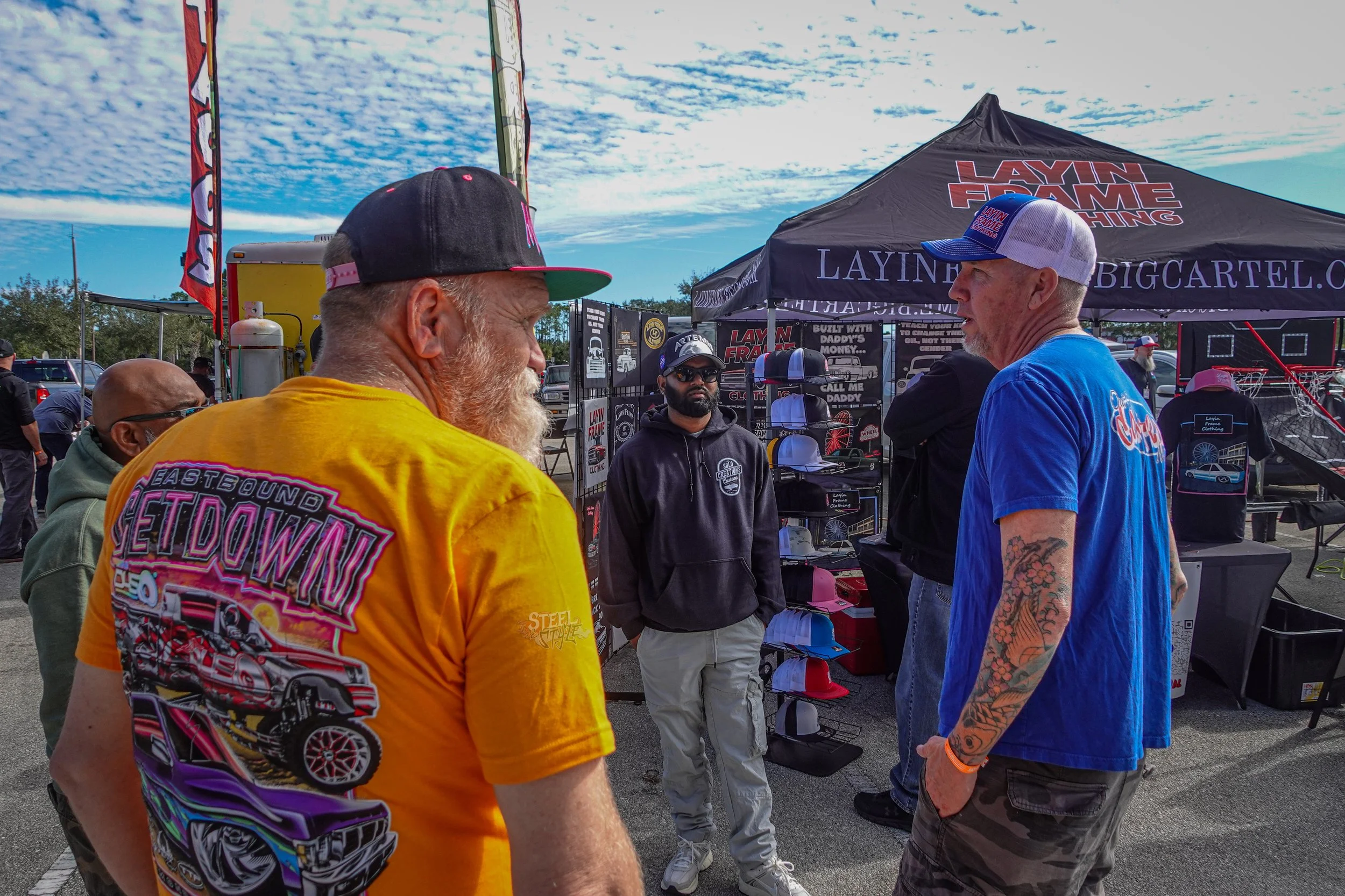 Group of men talking at an outdoor event with a vendor booth and tents in the background.