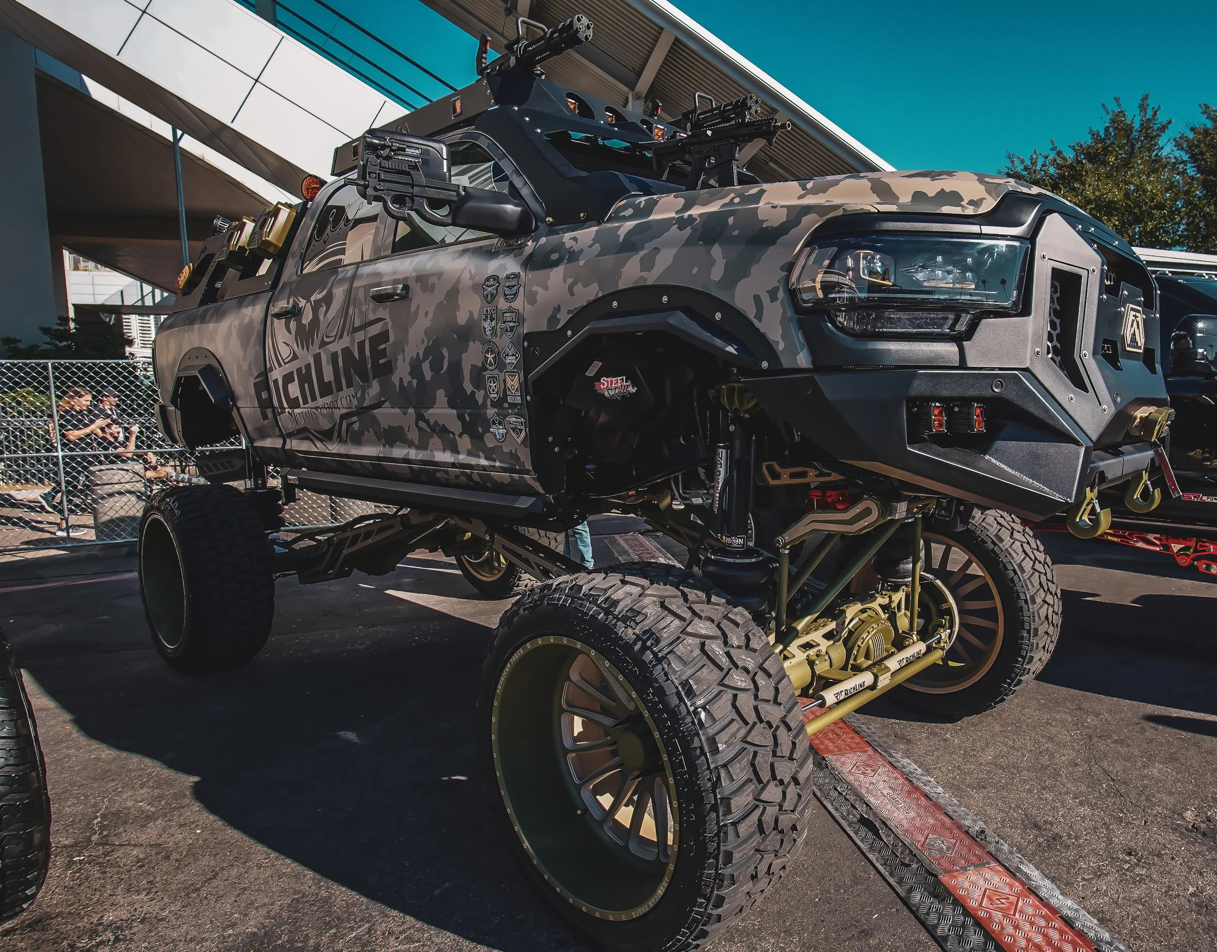 A heavily modified off-road vehicle with a camouflage paint job, large tires, and tactical equipment on the roof and sides, parked outdoors.