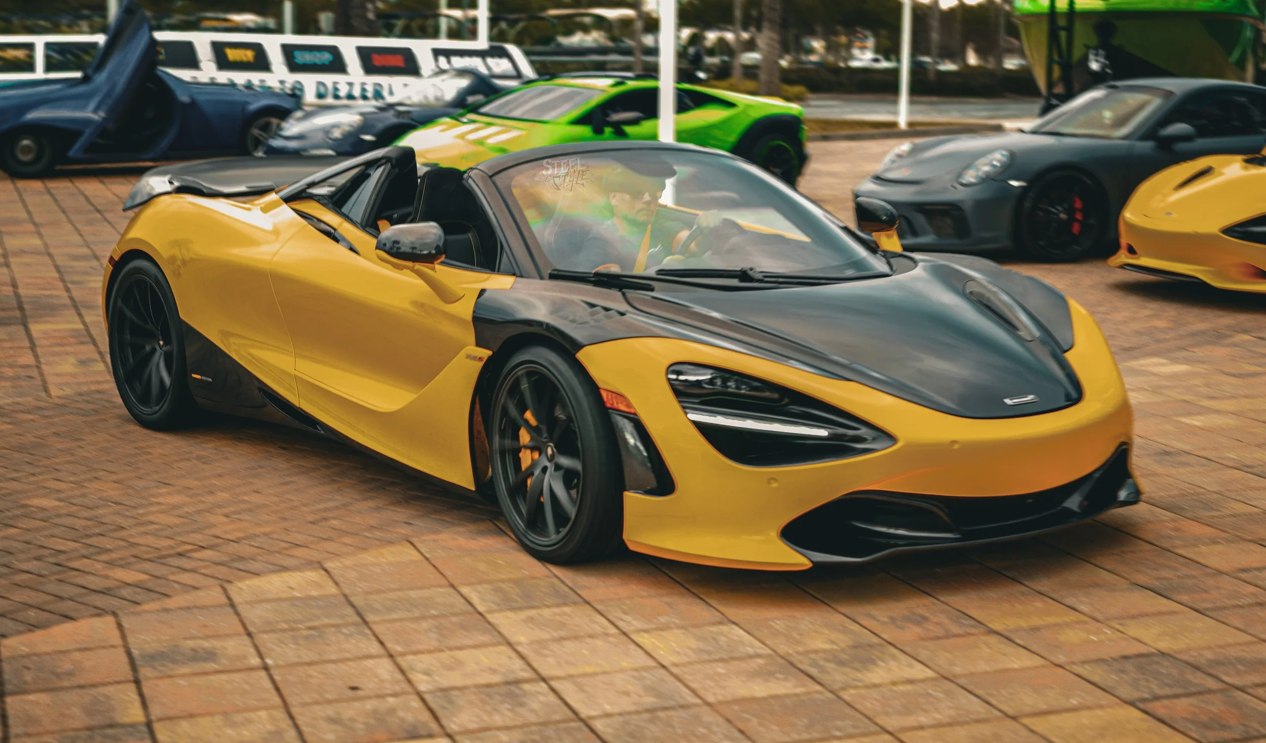 A yellow and black convertible sports car parked on a brick pavement, with several other luxury cars in the background.