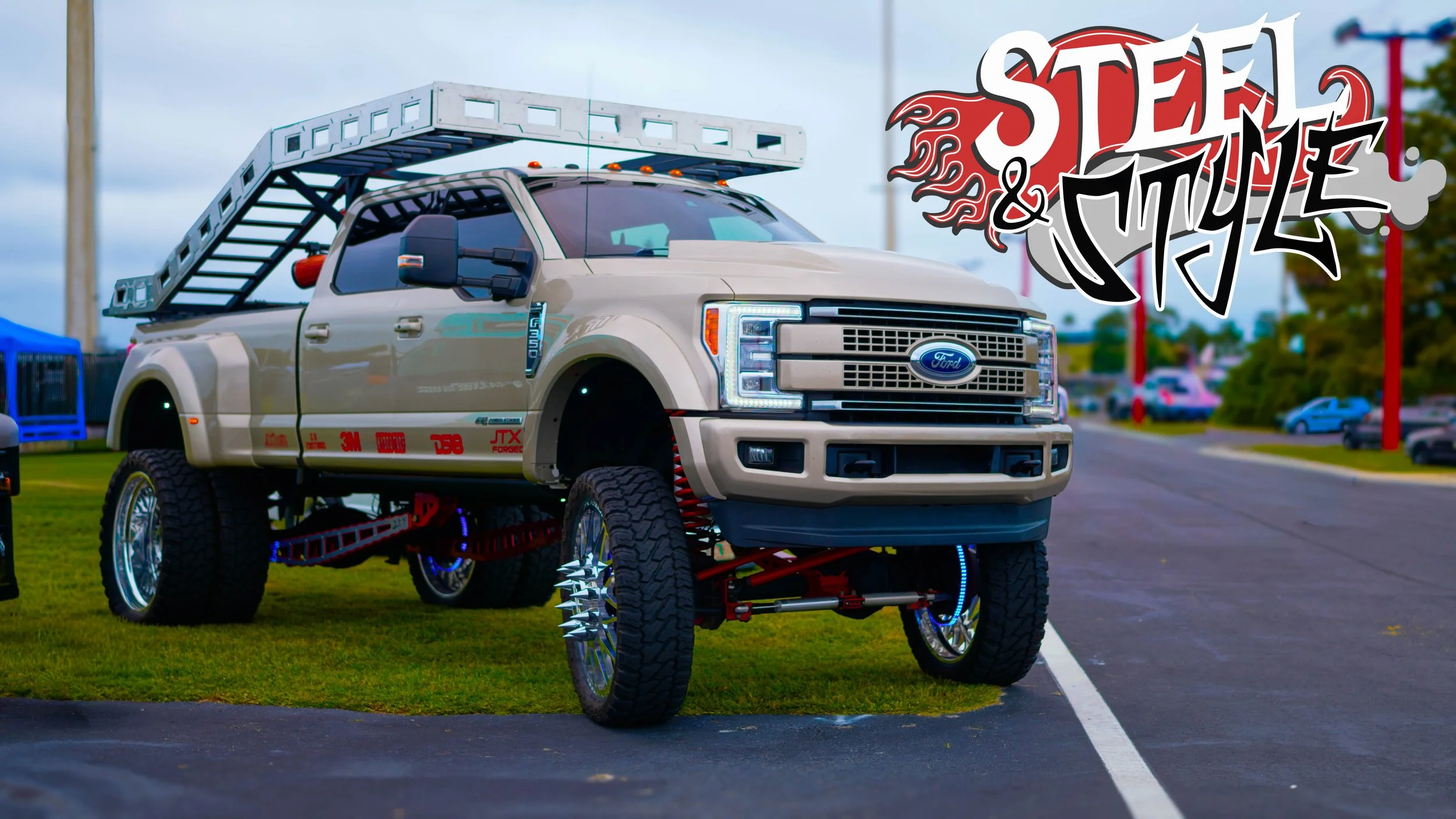 A modified Ford truck with oversized wheels and a ladder on top, parked on grass and pavement, featuring a 'Steam & Style' logo in the background.