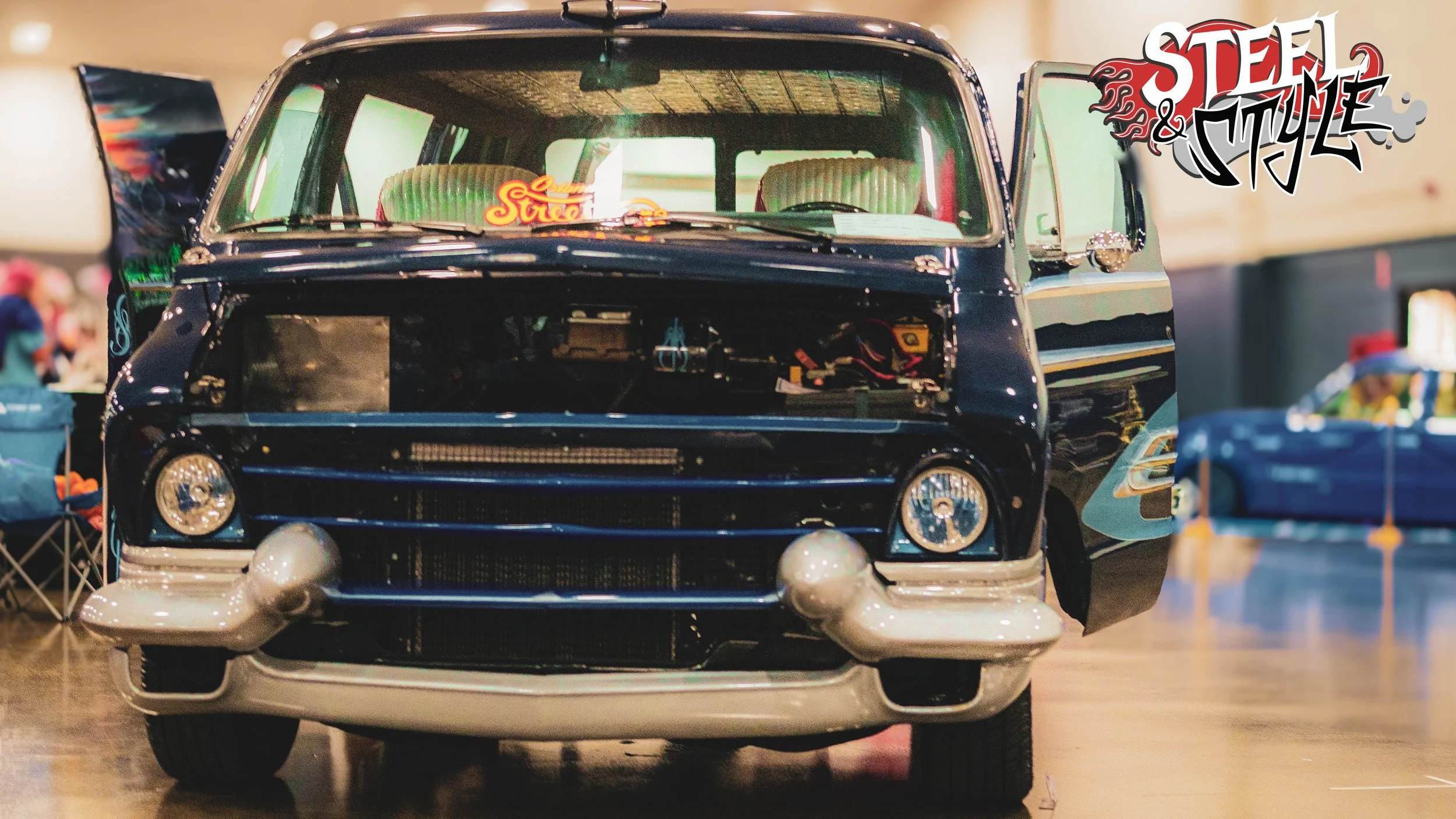 A vintage black van on display indoors, with part of its front and open rear doors exposed, showing the interior and engine components. The van has round headlights, a chrome bumper, and a large windshield with a neon sign inside reading 'Street'. Th