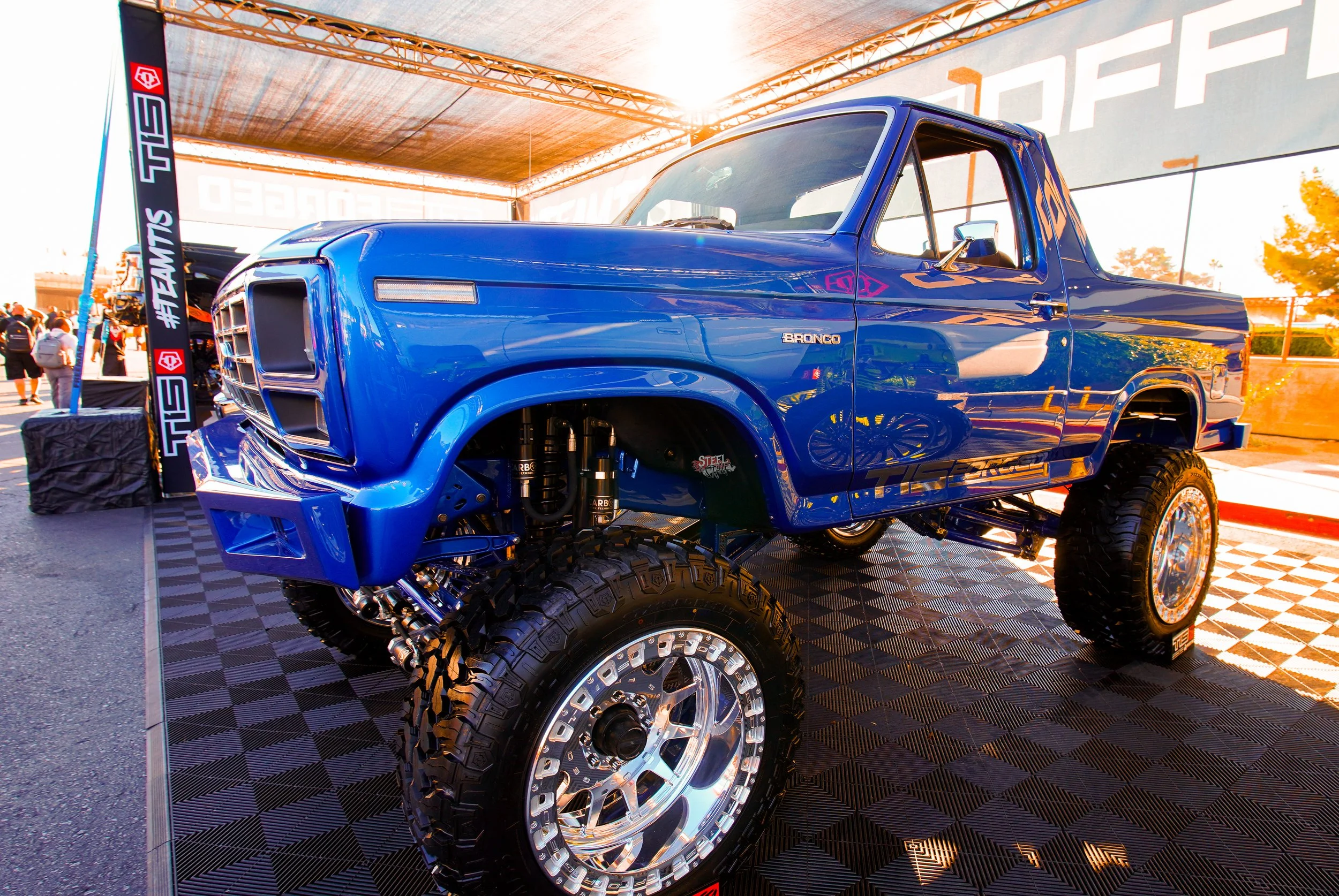 A custom blue Ford Bronco truck with large off-road tires and suspension on display at an automobile event under a canopy with people in the background.