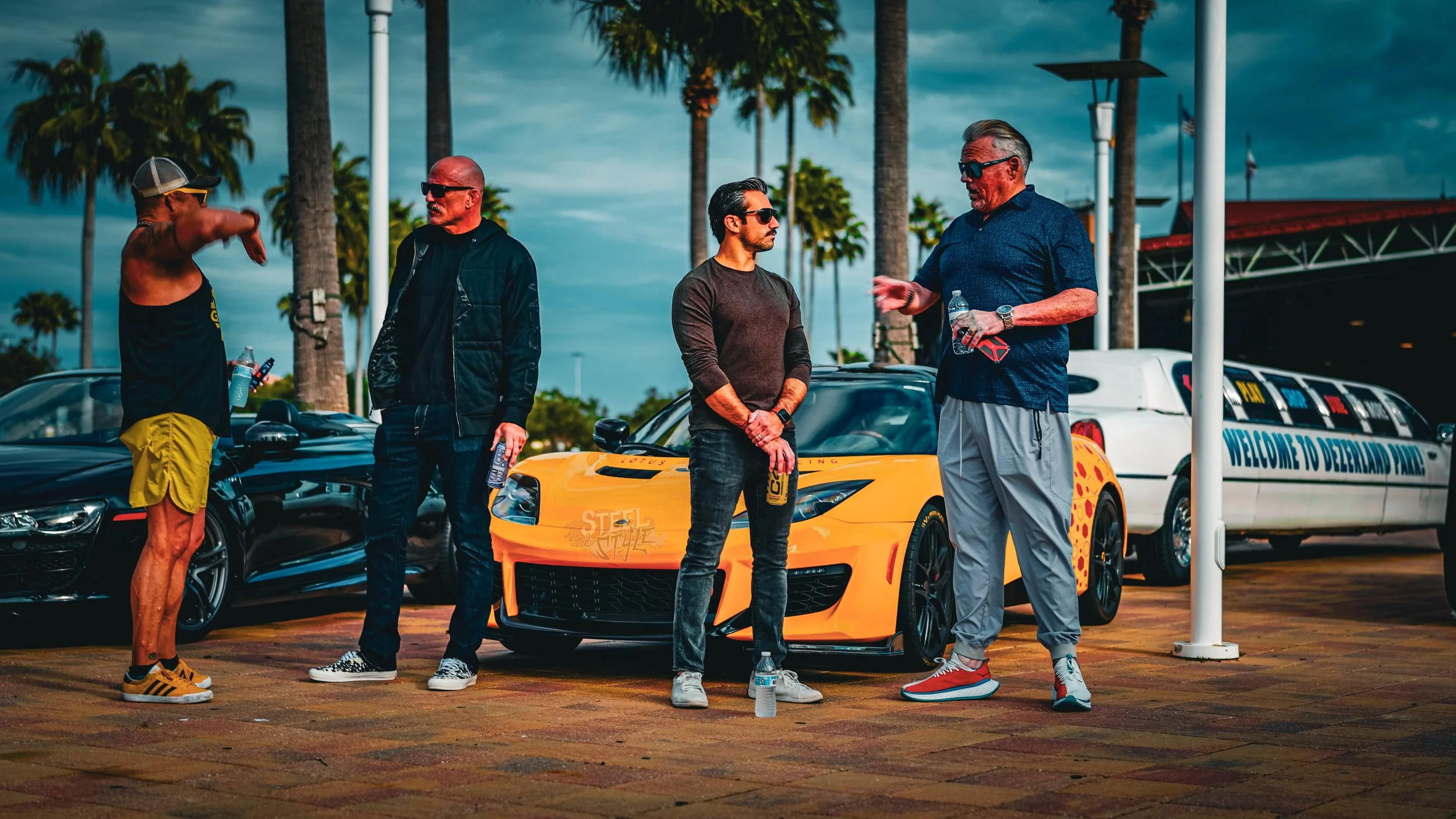 Four men standing near cars, including a yellow sports car, at a race track or car event on a cloudy day with palm trees in the background. They are engaged in conversation, holding water bottles.