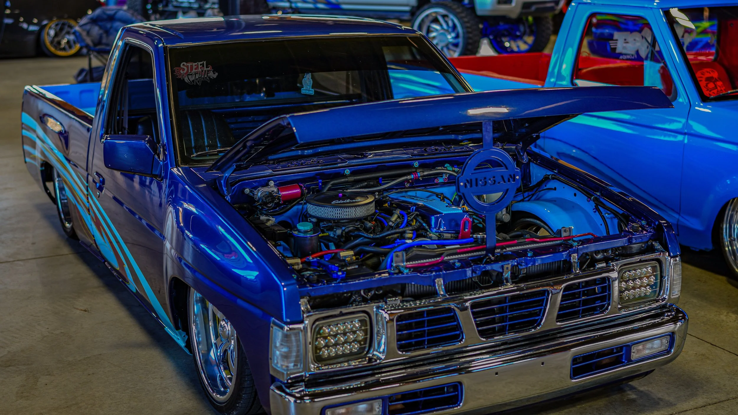 Blue customized Nissan pickup truck with its hood open, showcasing its engine at an indoor car show.