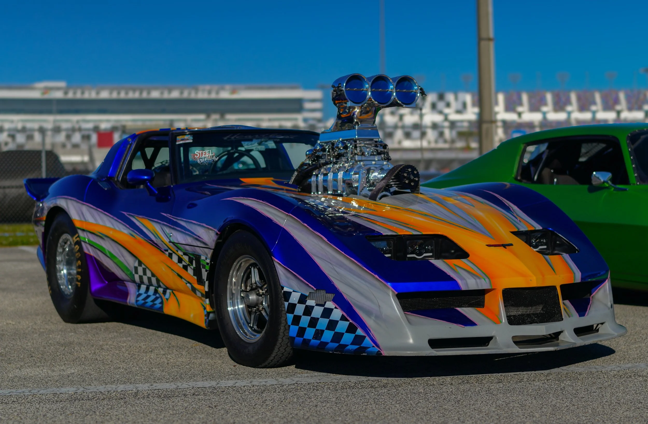 Colorful race car with a large exposed supercharger on the hood, parked next to a green car on a race track.