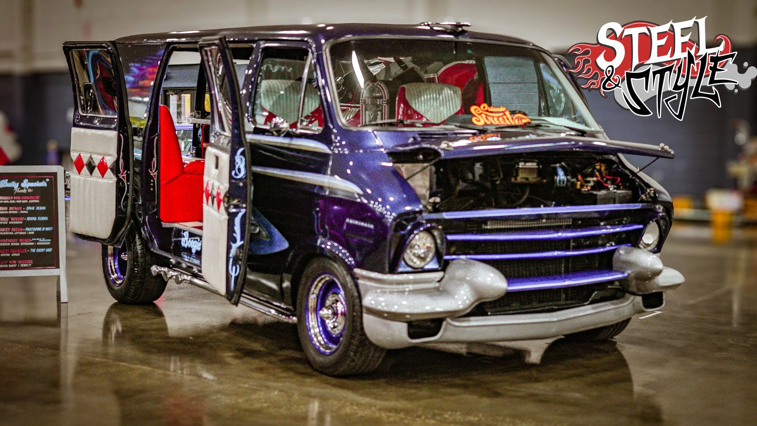 A customized vintage car with a dark blue body, red interior seats, and one door open on display at an indoor event, with a 'Steel & Style' logo in the upper right corner.