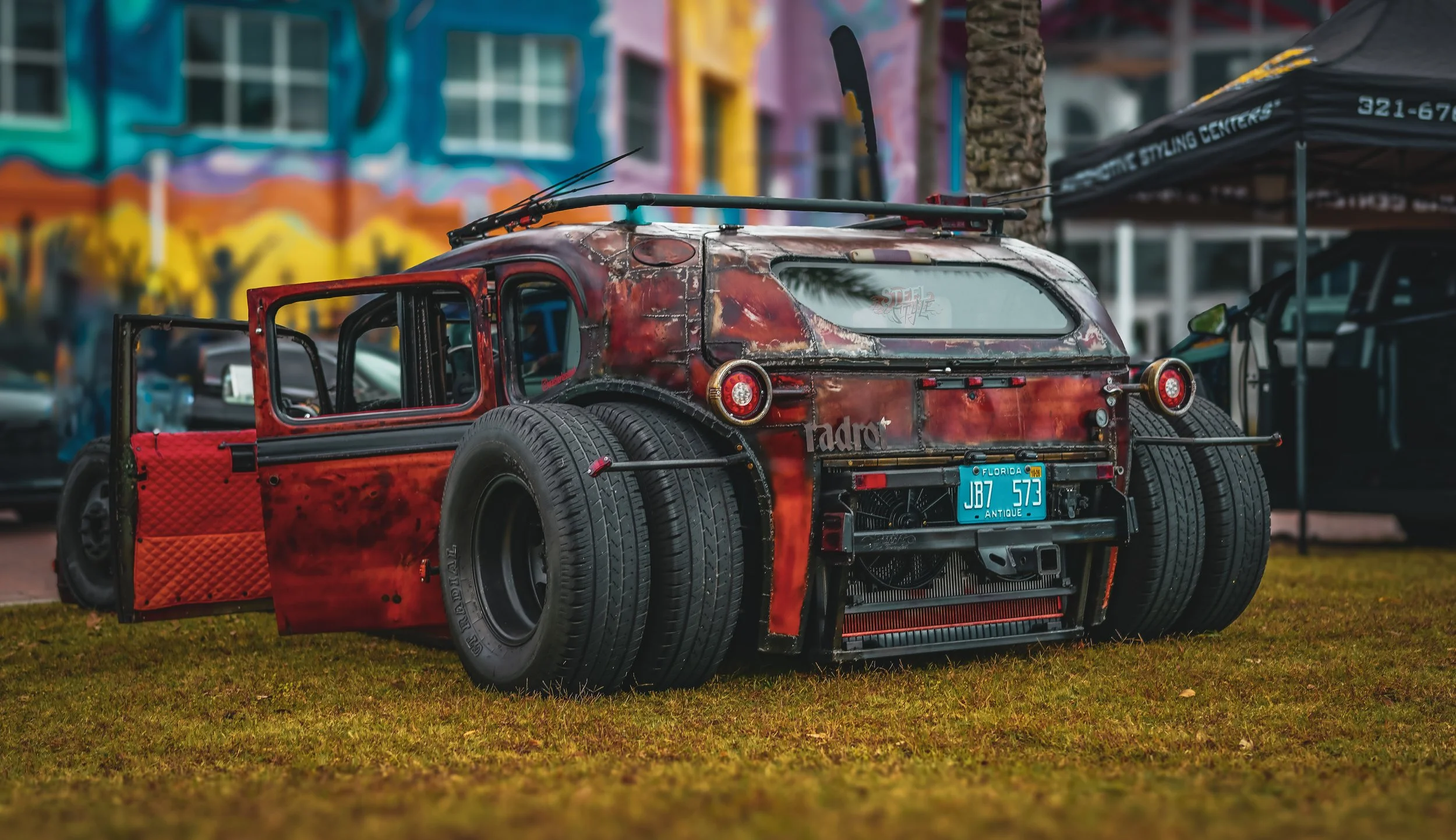 A custom-built, vintage-style car with a distressed red paint job, large rear tires, and a Florida antique license plate, displayed outdoors on grass with a colorful building and tents in the background.