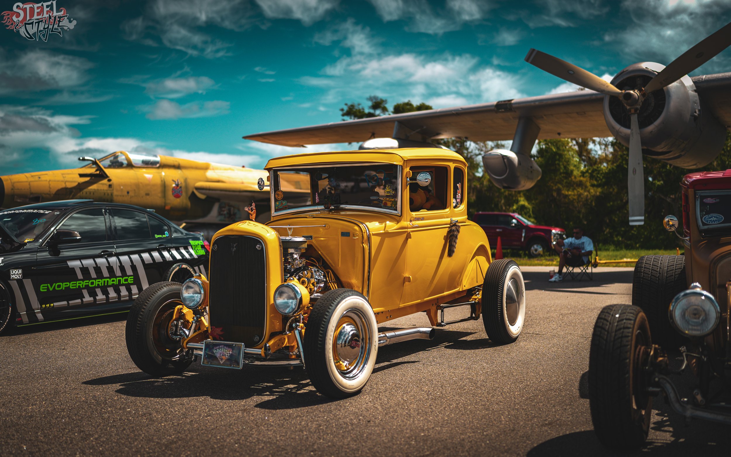 A vintage yellow hot rod car is displayed among other vehicles at an outdoor car show, with a propeller airplane and a black race car nearby and spectators in the background.