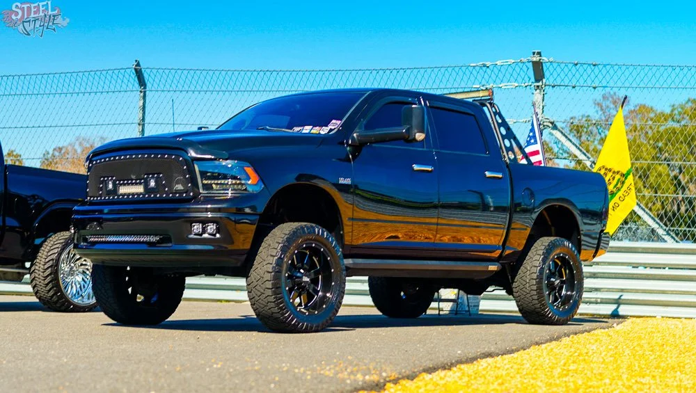 A black lifted pickup truck with off-road tires and custom wheels parked in front of a chain-link fence at a race track, with flags waving in the background.