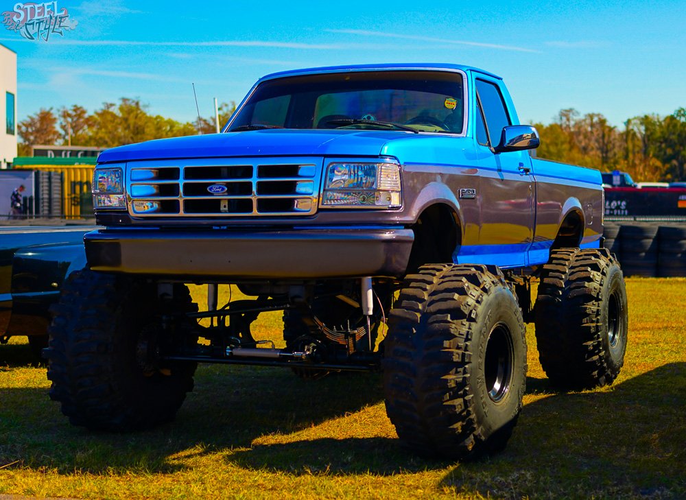 A blue Ford pickup truck with large off-road tires and lifted suspension, parked on grass at an outdoor event.
