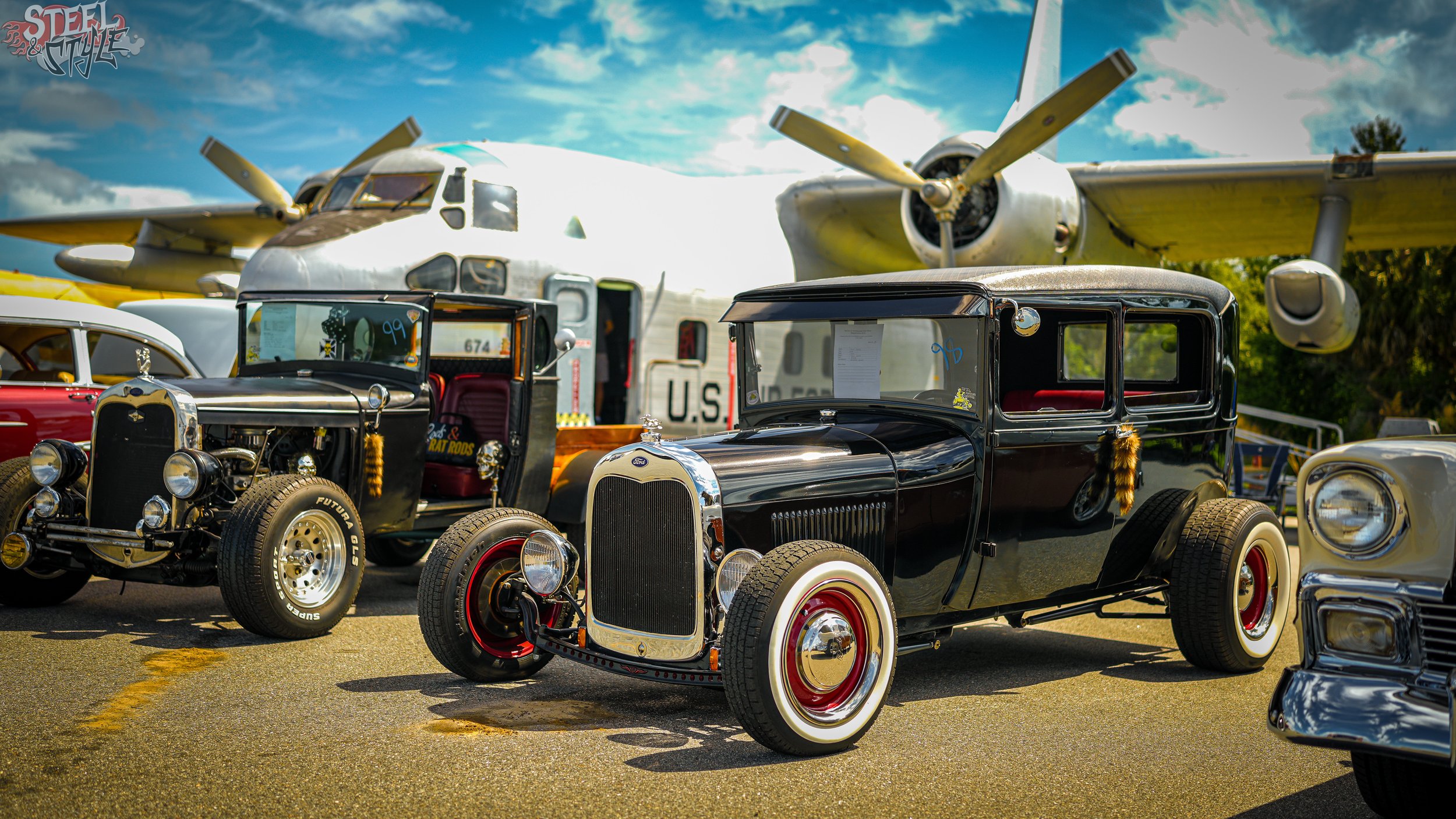 Vintage cars displayed outdoors with a large airplane in the background against a partly cloudy sky.
