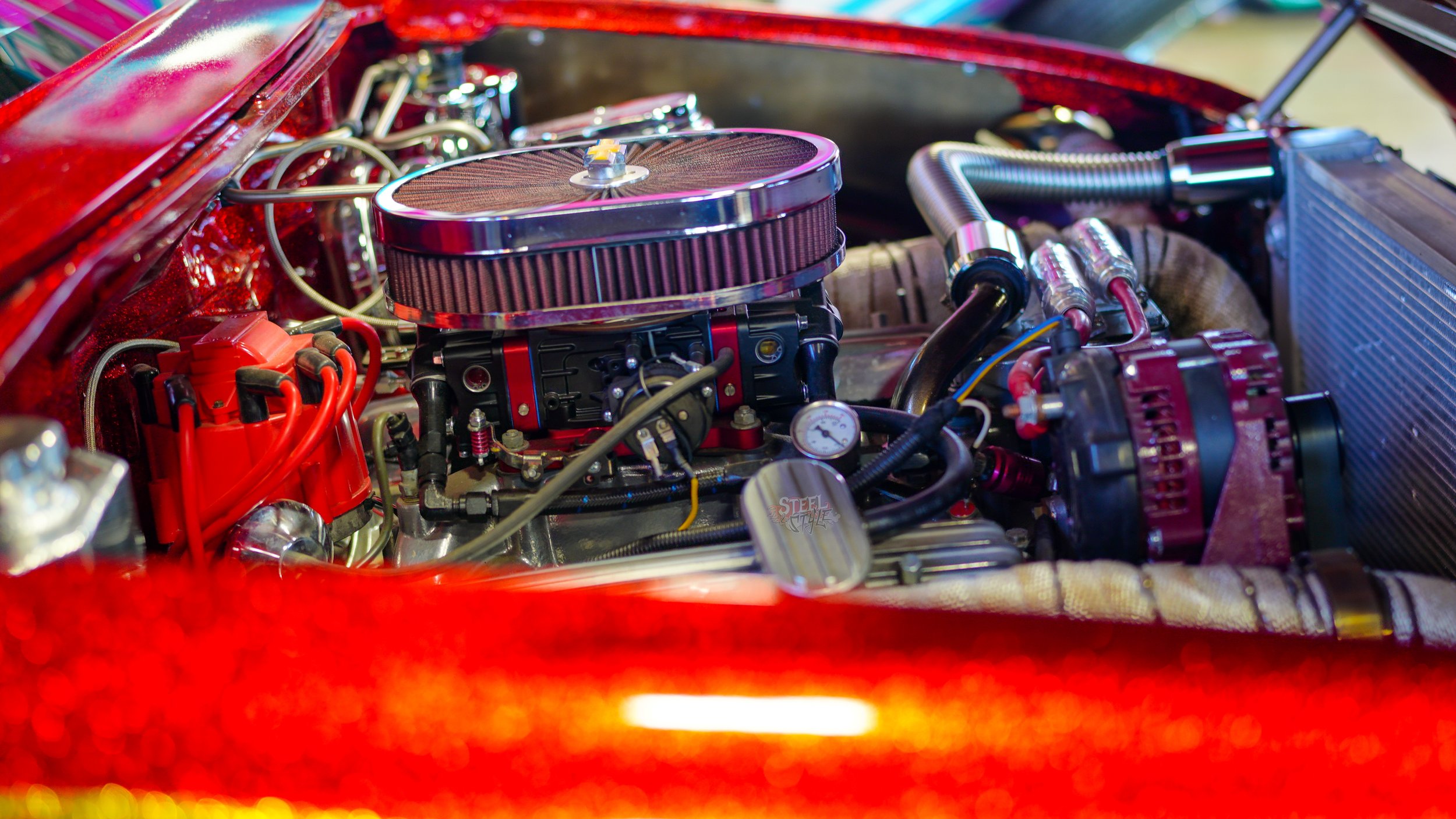 Close-up view of a high-performance car engine with a chrome air filter, red distributor cap, and various metallic components, surrounded by a red-painted engine bay.