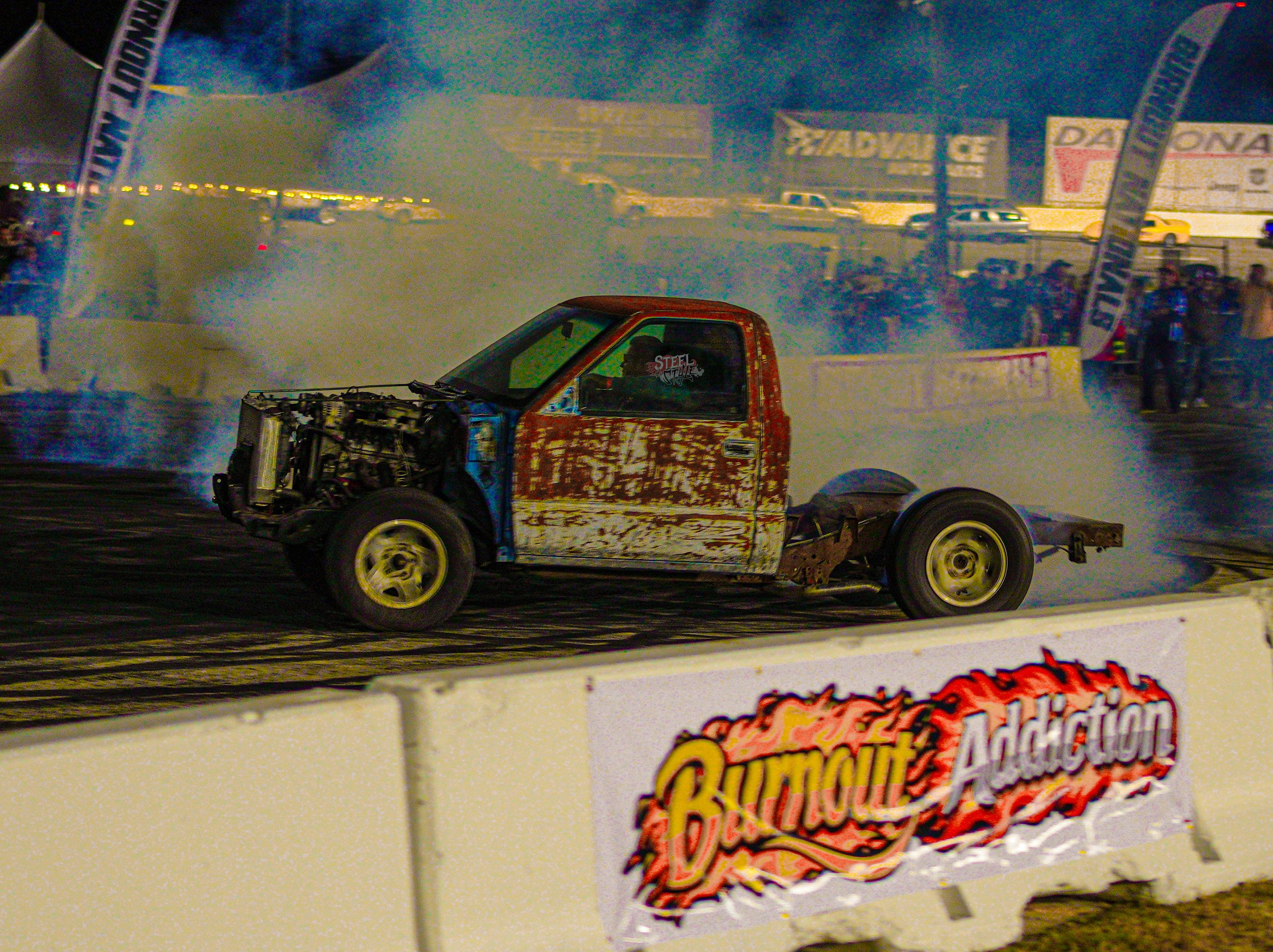 A rusty, stripped-down race car on a drag strip with smoke and tire marks, with spectators and banners in the background.