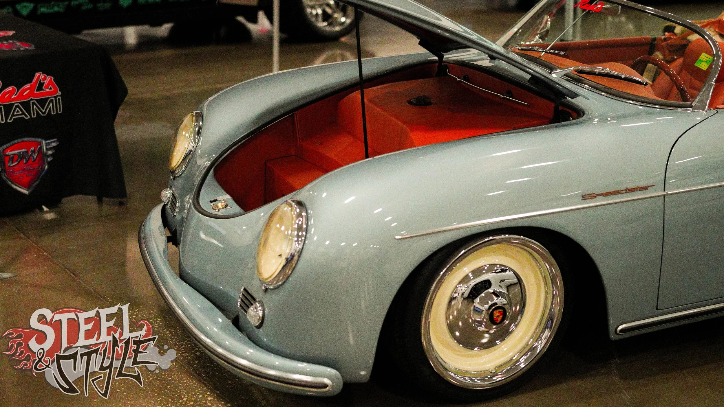 A vintage light blue Porsche Speedster with its front trunk open, revealing a red interior. The vehicle is displayed at an indoor car show, with a Steel & Style logo visible on the floor near the vehicle.