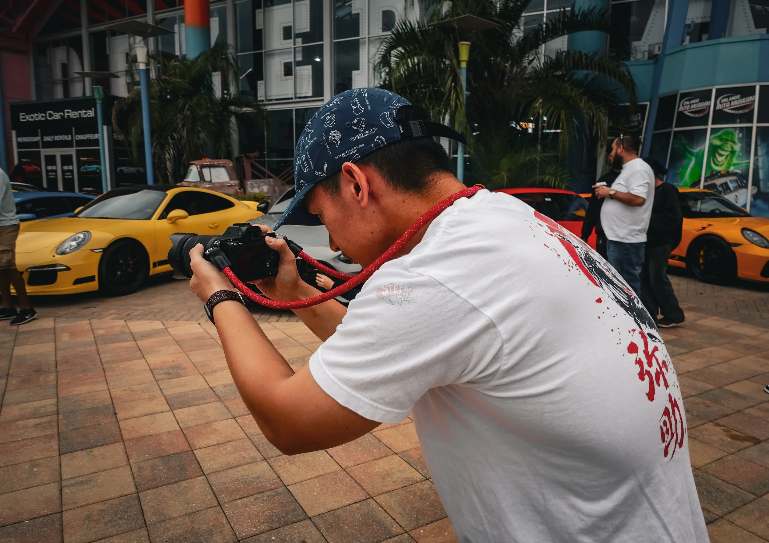 Young man taking a photograph with a camera at a luxury car event, with yellow sports cars and people in the background.