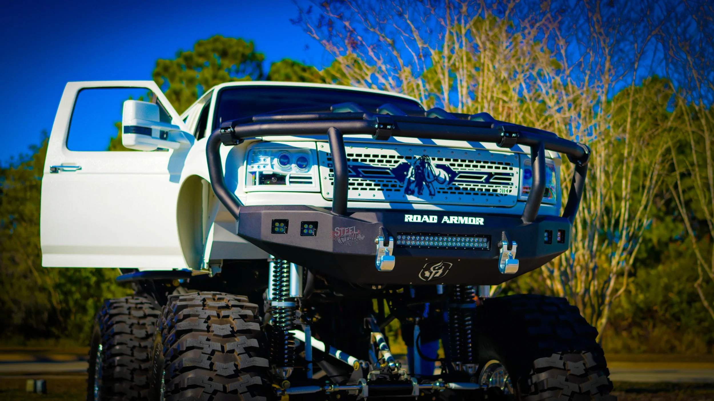 A custom off-road vehicle with large tires, a reinforced front bumper, and a grill guard, parked outdoors under a blue sky with trees in the background.