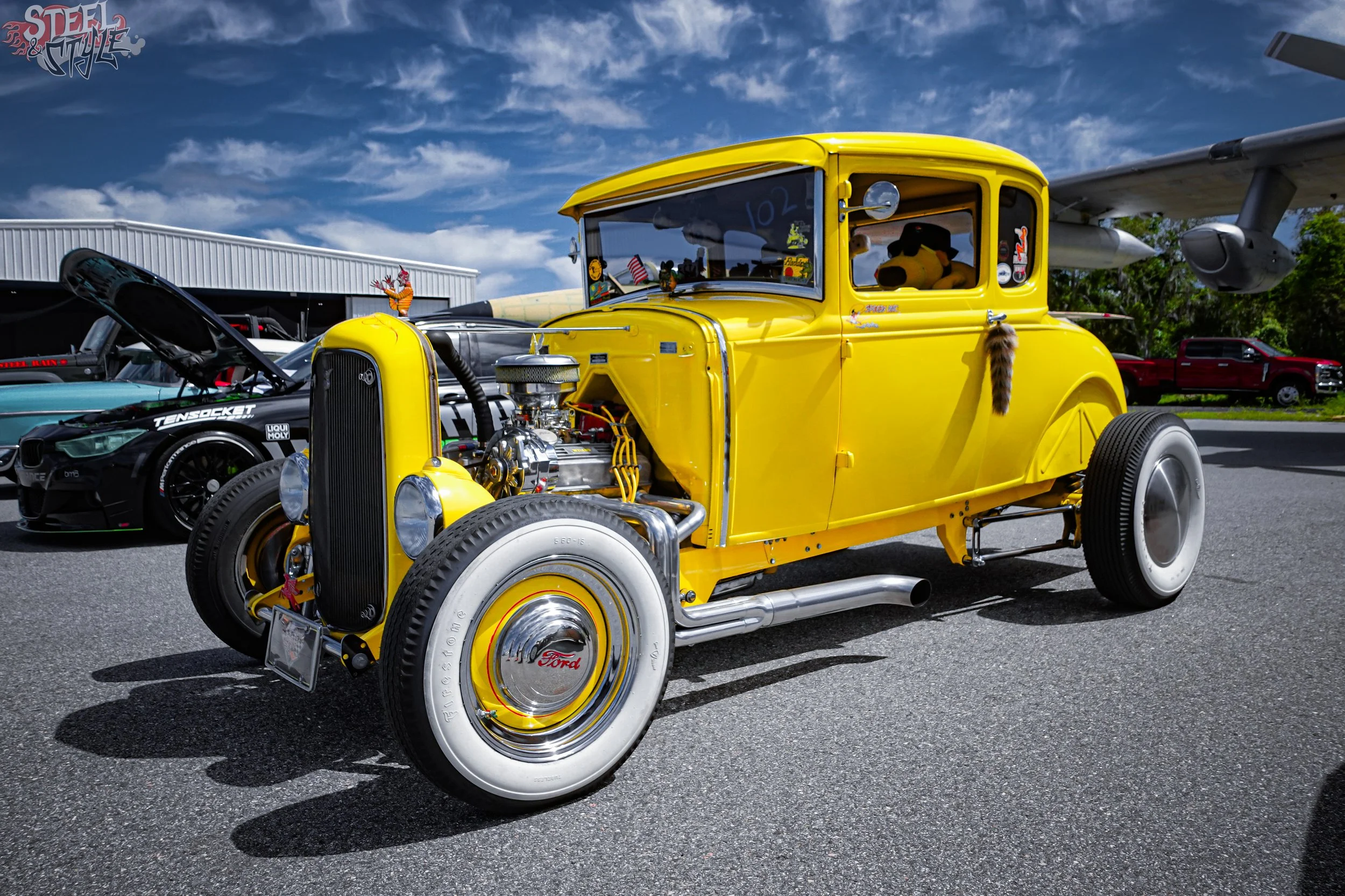 A vintage yellow Ford hot rod with an exposed engine and white wall tires parked outdoors, next to a modern black sports car with a racing decal, under a blue sky with clouds and a building in the background.