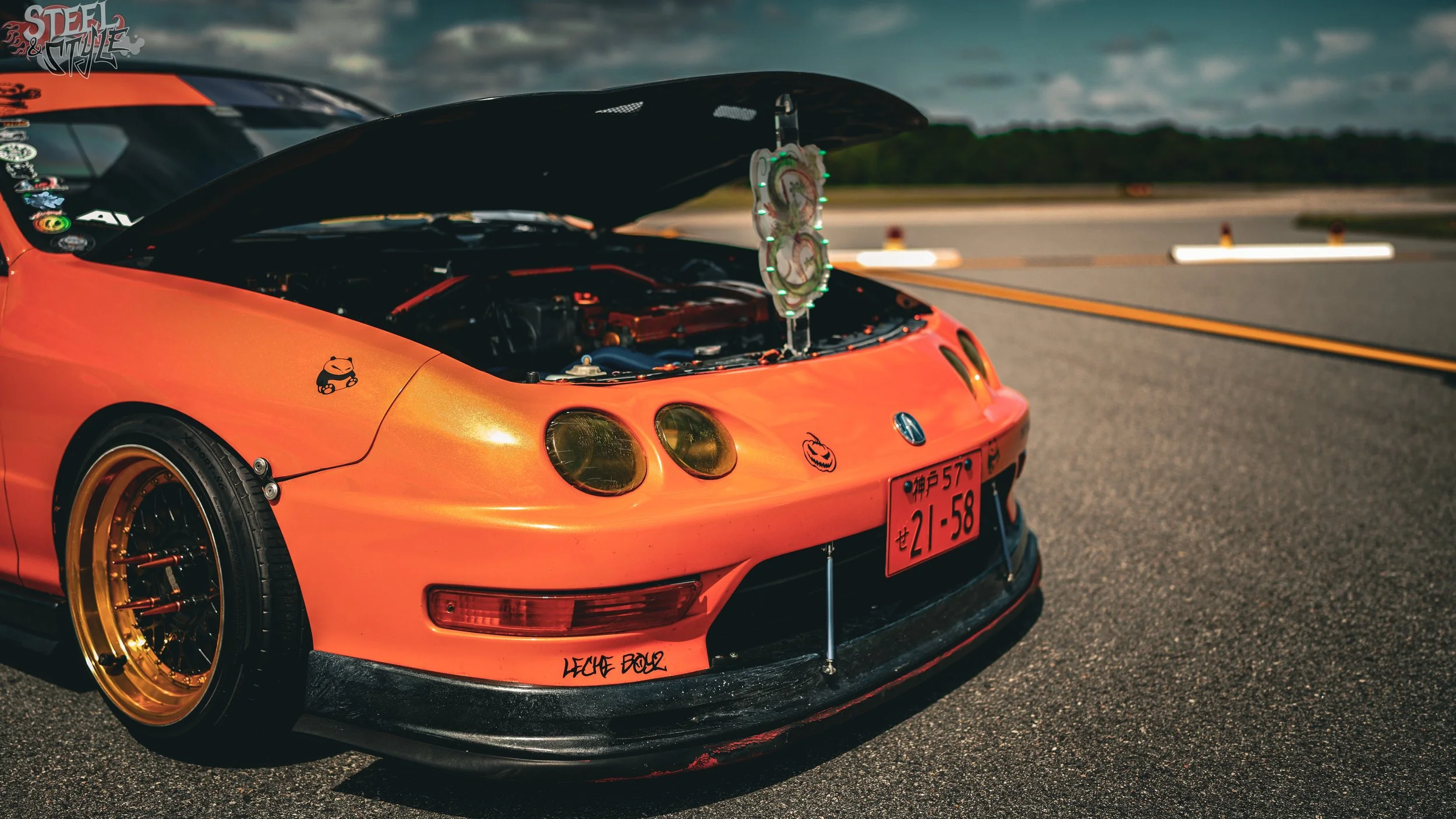 An orange sports car with its hood open shows two turbochargers with glowing green lights in a parking lot during overcast weather. The car has custom black and gold wheels, stickers, and a Halloween-themed pumpkin decal on the front.