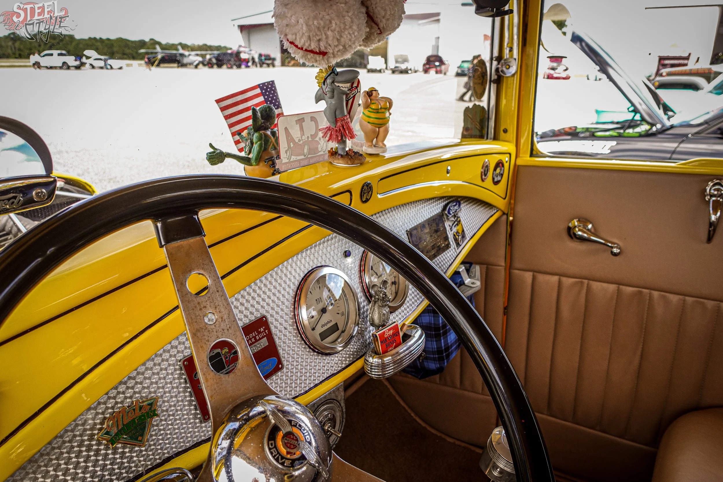 View from inside a vintage yellow car with a dashboard decorated with various small figurines, including a shark, an alligator, a woman in a swimsuit, and a small American flag, parked in a lot with other vehicles outside a large building.