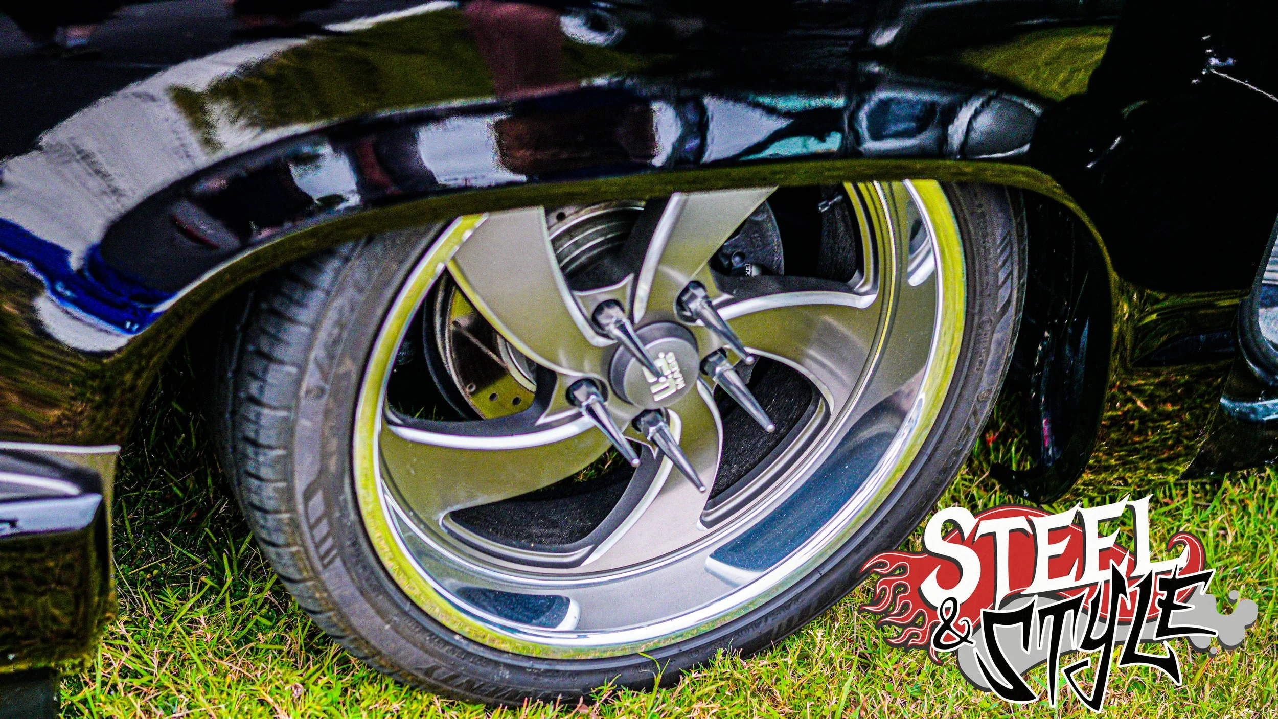 Close-up of a car's chrome wheel and black tire on grass with a 'Steel & Style' logo in the bottom right corner.